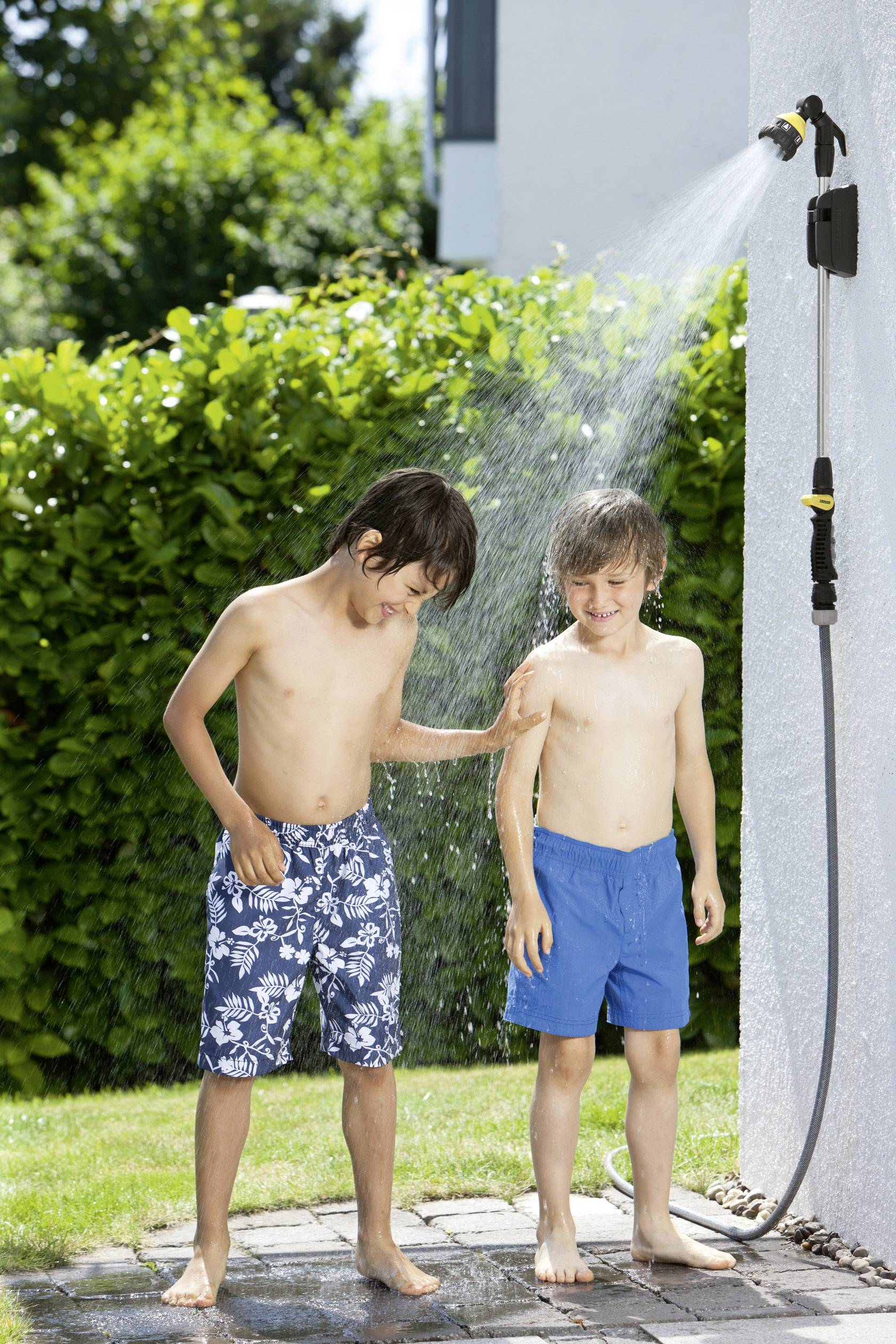 Two boys are playing beside an outdoor shower in the garden. One is spraying the other with water. It is a sunny day.