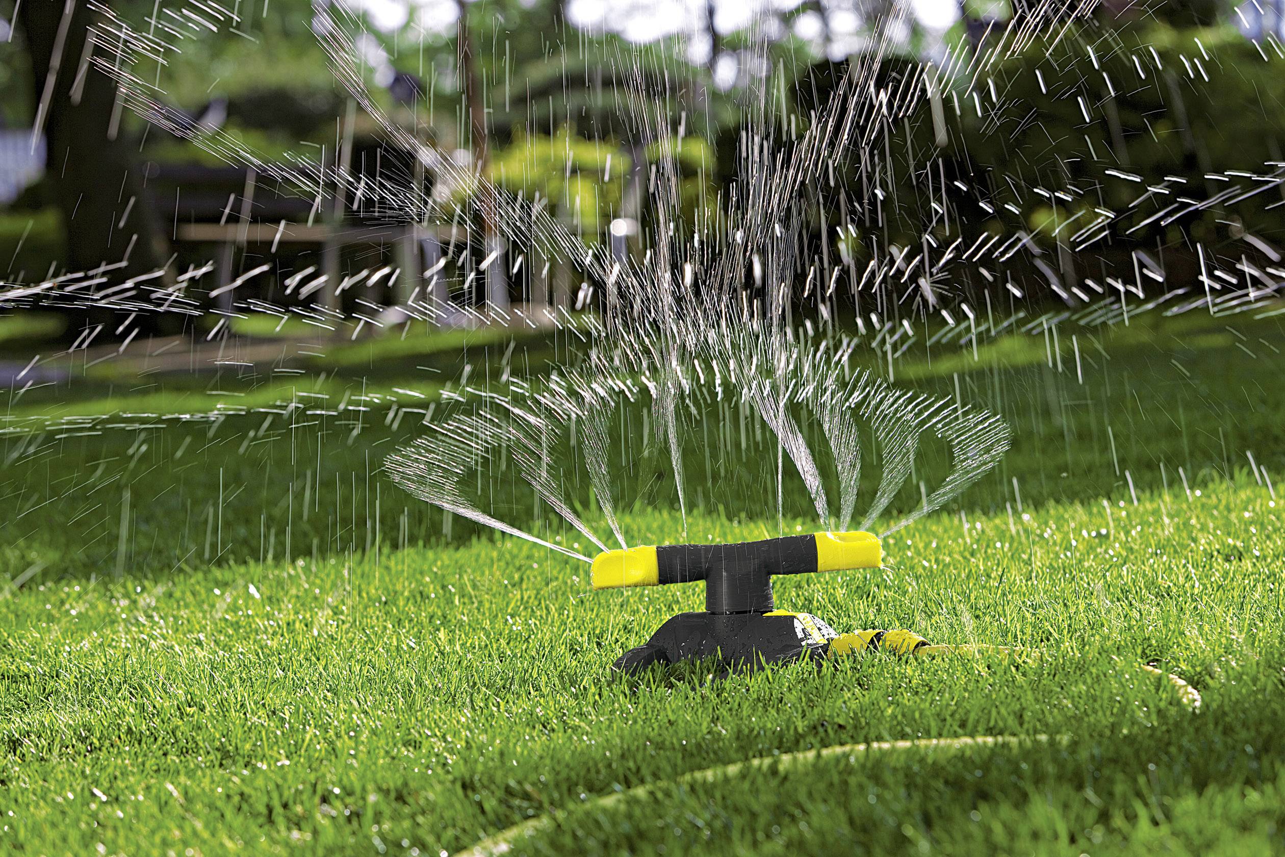 A lawn sprinkler on a green lawn distributes water in circular patterns. The background shows blurred trees and benches.