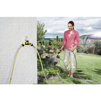 A person is watering flowers with a garden hose in a garden. A distributor is attached to the outside water tap. Lawn and bushes are visible in the background.