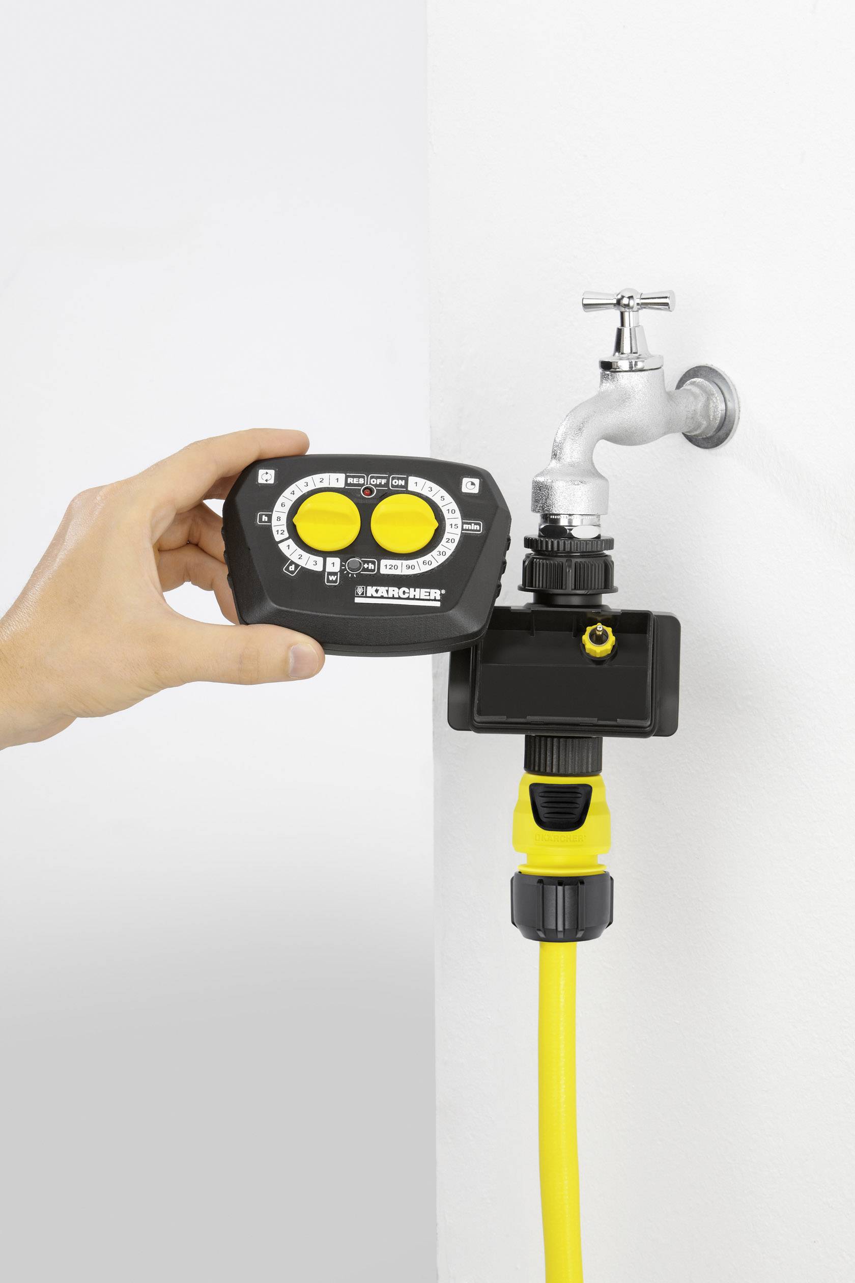 A hand is holding an irrigation control device in front of a tap with a yellow hose connector, ready for installation.