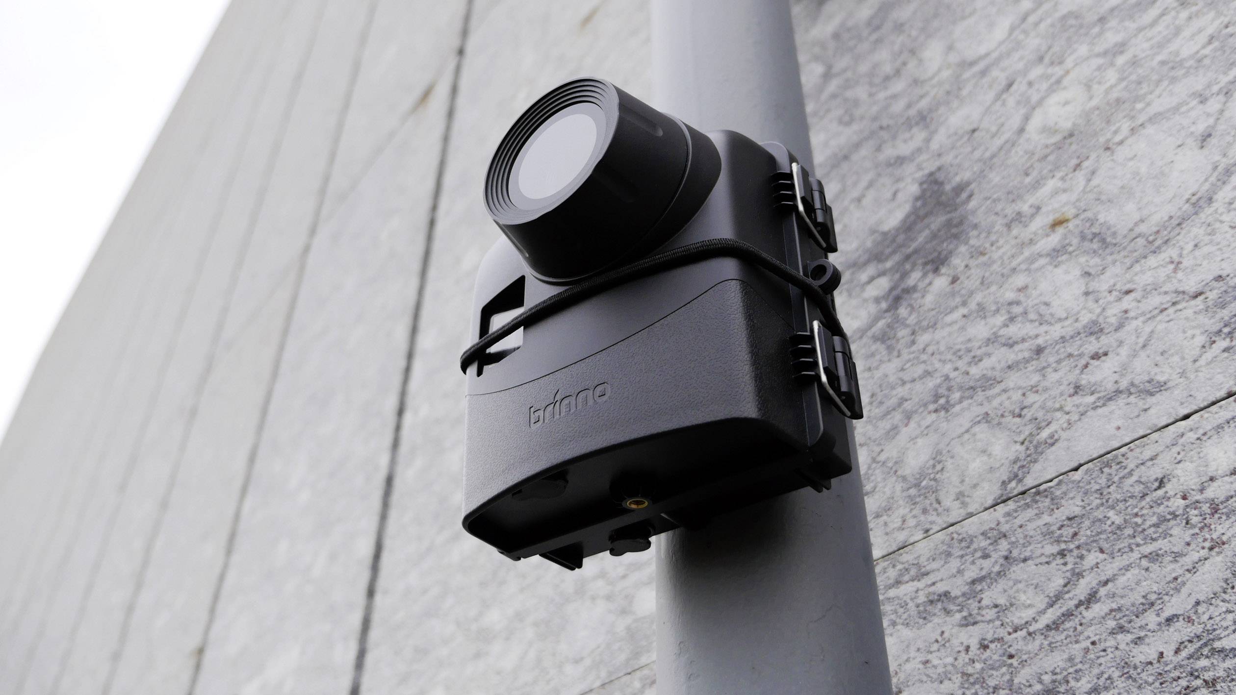 A black security camera is mounted on a grey pole and attached to a stone wall. The camera is facing left.