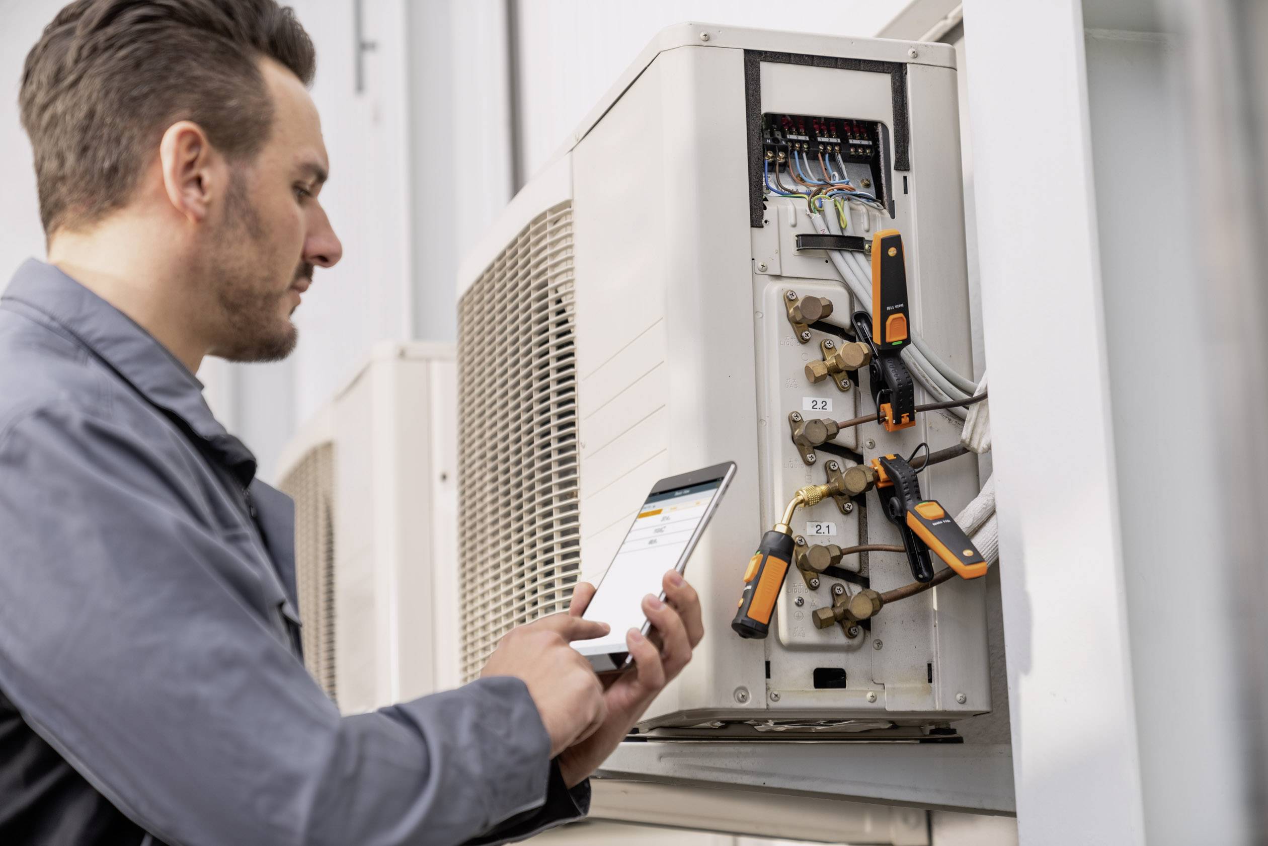 A technician is checking an air conditioning unit with a tablet. The system has several connected hoses and measuring instruments.