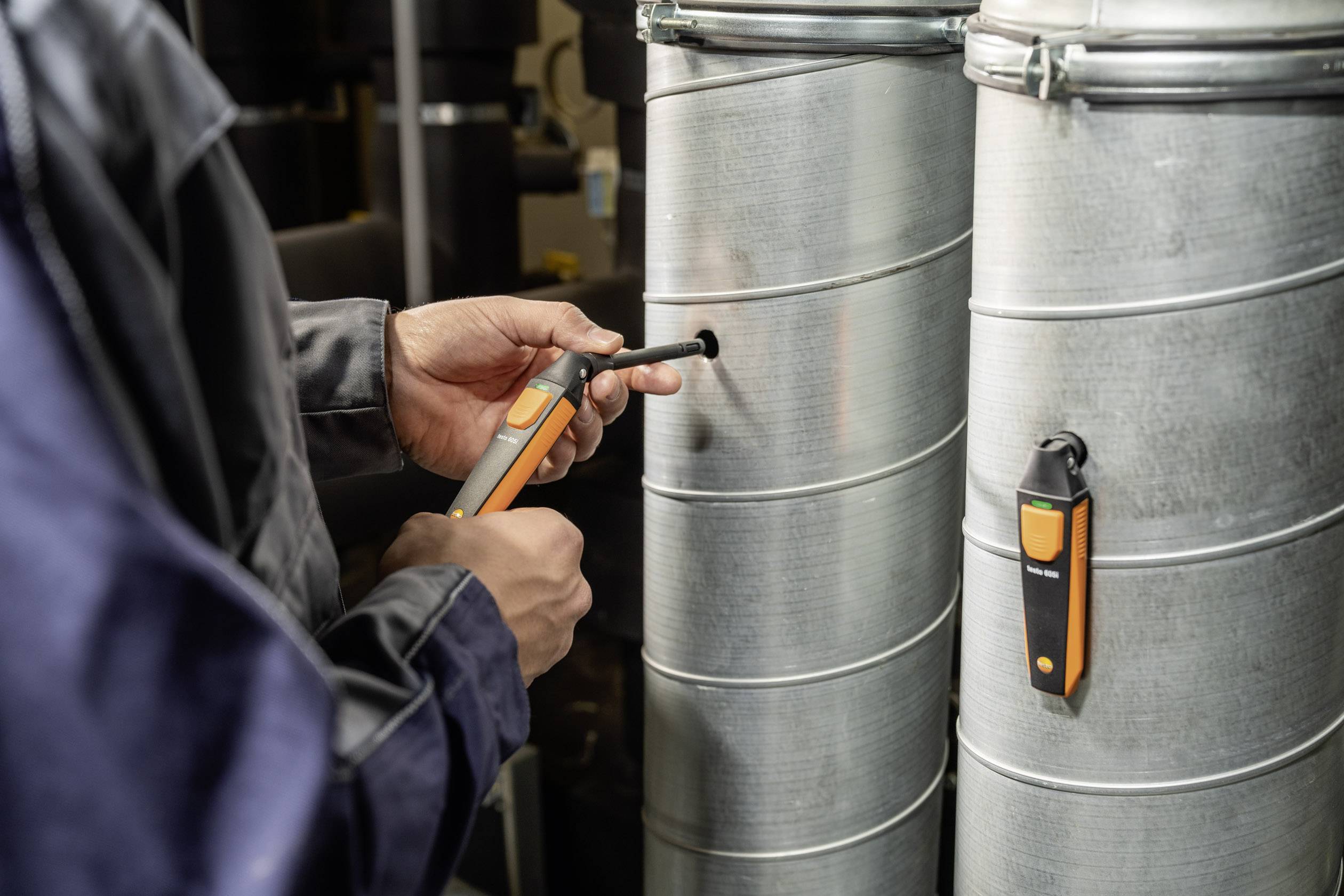 Close-up of a person in work attire using a temperature measurement device on a metallic ventilation pipe.