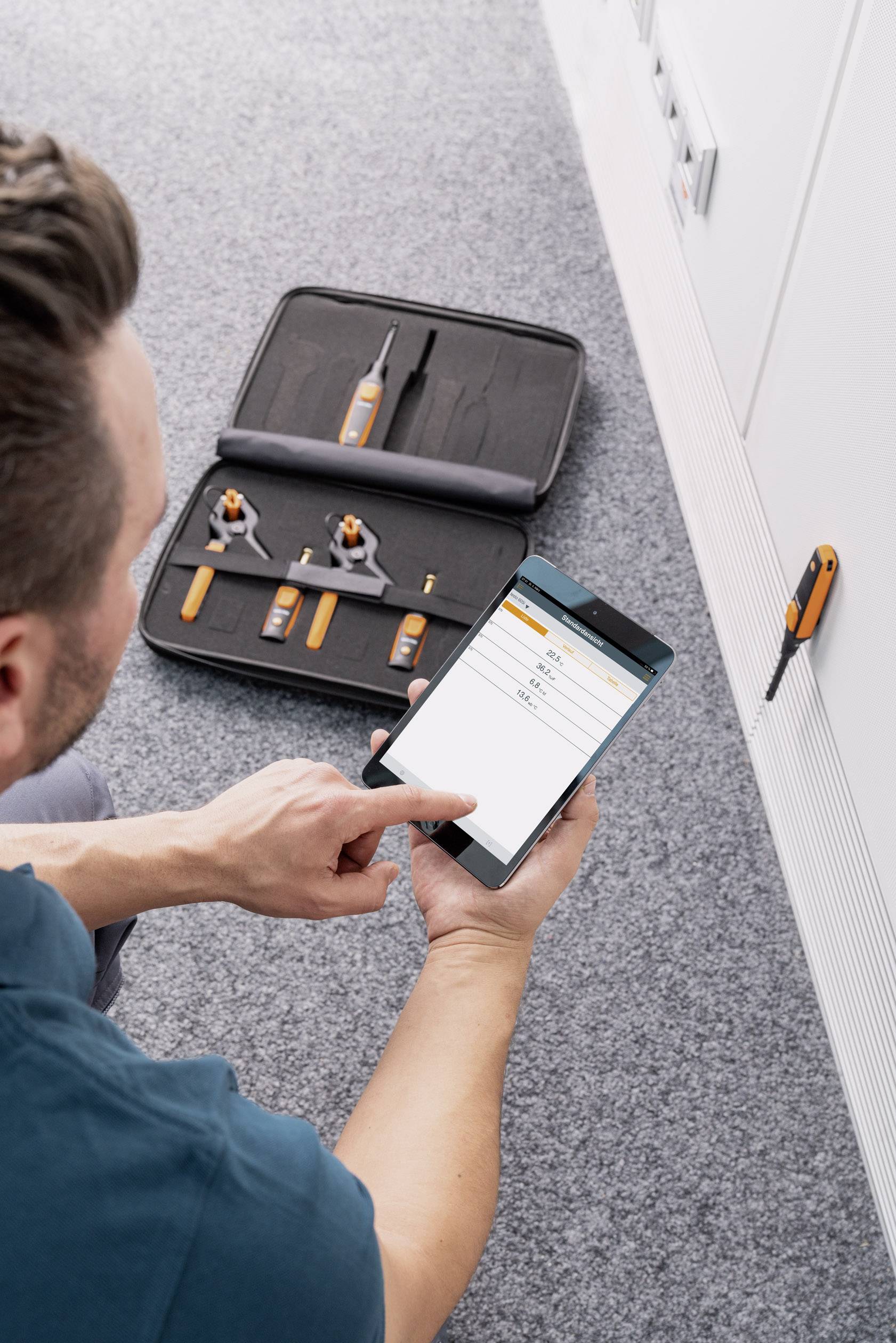 A man is kneeling on a carpet, operating a smartphone app to control an assembly process. In the background, an opened toolset is lying nearby.