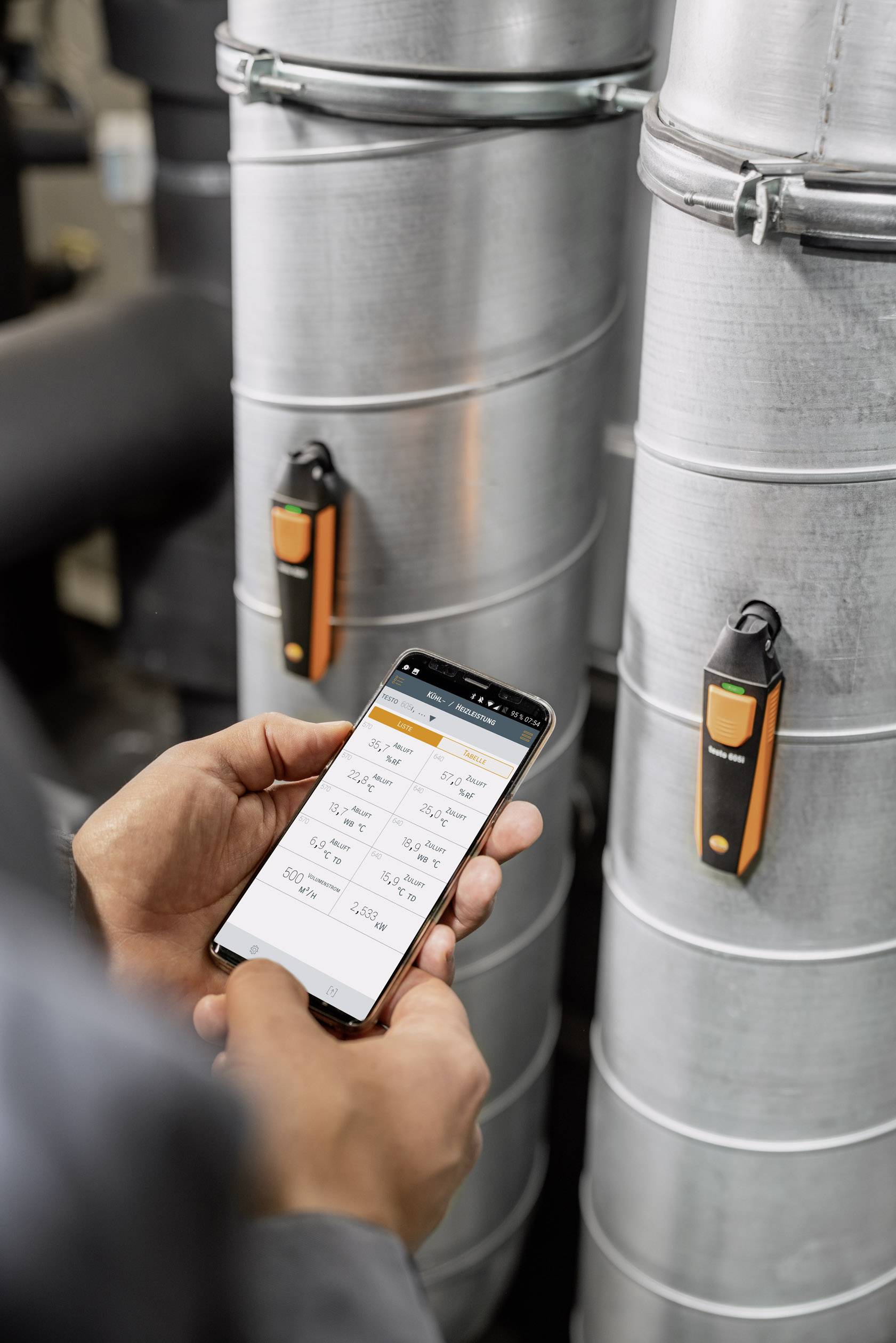 A person is using a smartphone to monitor data on two silver pipes with orange sensors.