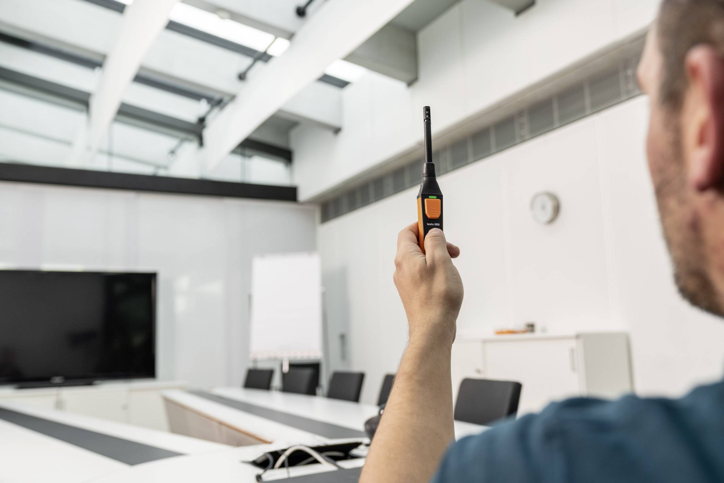 A person is holding a measuring device in a conference room, presumably to measure air quality or temperature. In the background, there is a large screen and a whiteboard.