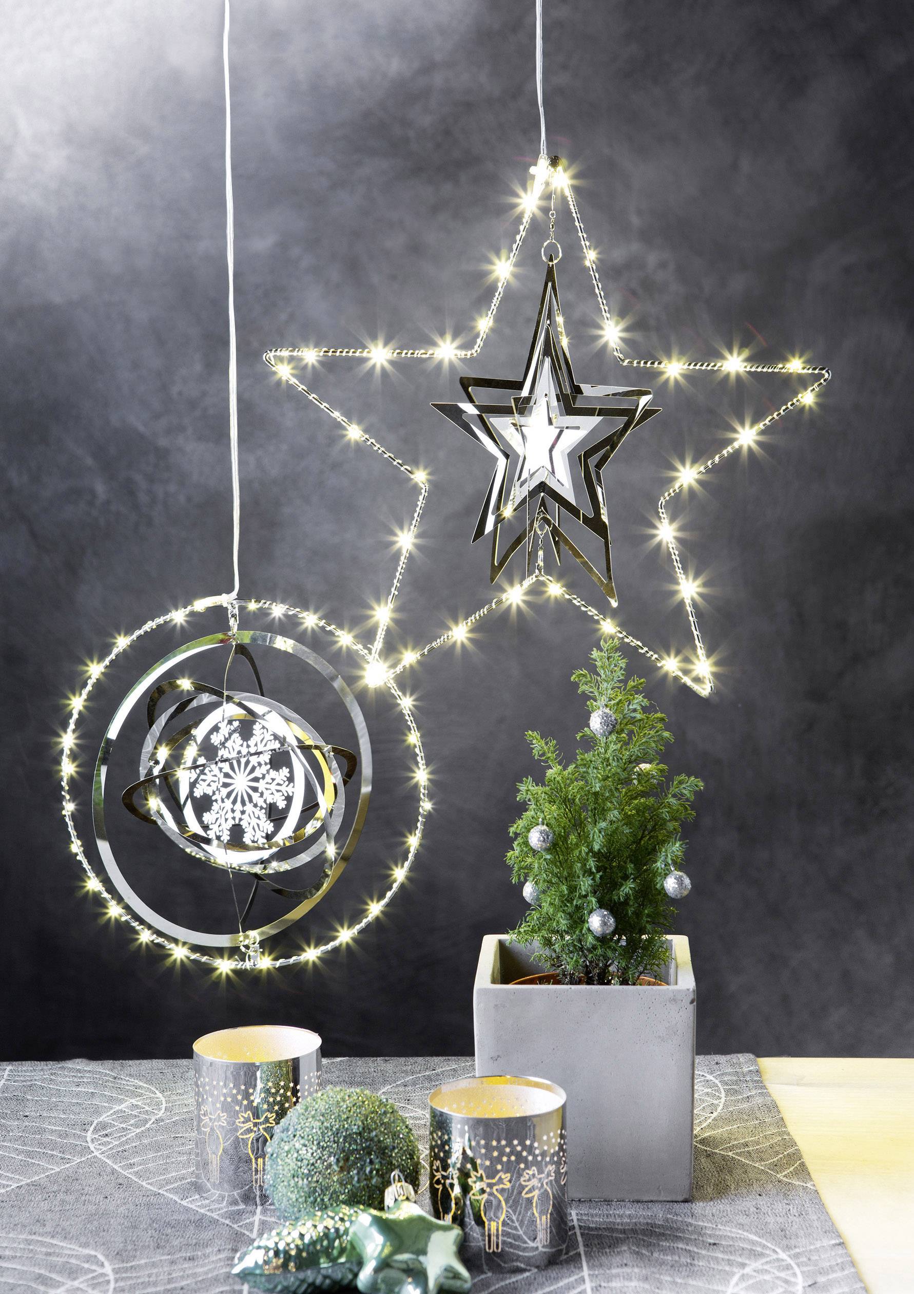 A small, decorated Christmas tree stands in a pot next to glowing star and snowflake ornaments. Two candles are placed in front of it.
