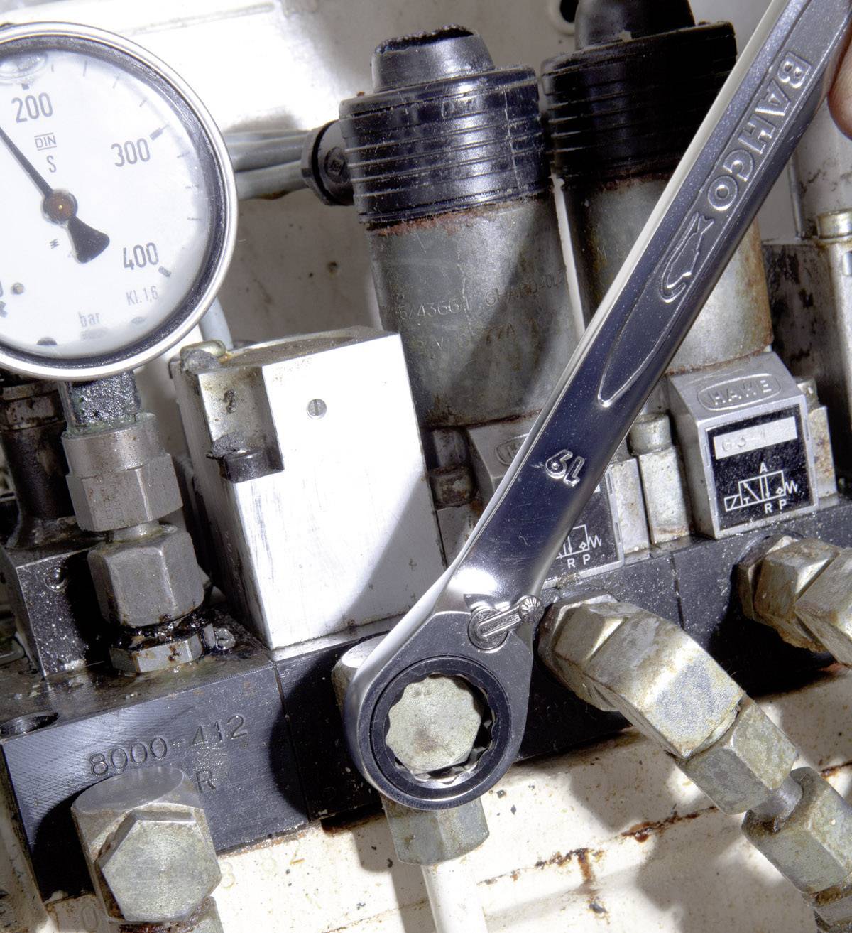 A spanner turns a screw on a machine component. A pressure gauge shows a reading of 200.