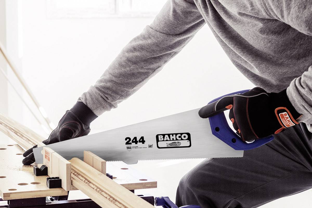 A person is sawing a wooden board on a workbench using a Bahco handsaw. The person is wearing gloves and a long-sleeved shirt.