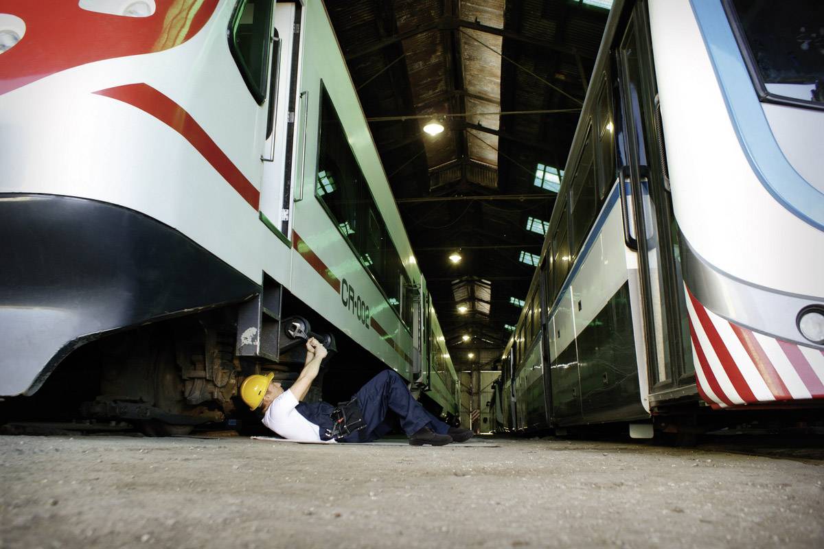 A mechanic is working underneath a stationary train in a workshop hall. Additional trains can be seen to the left and right.