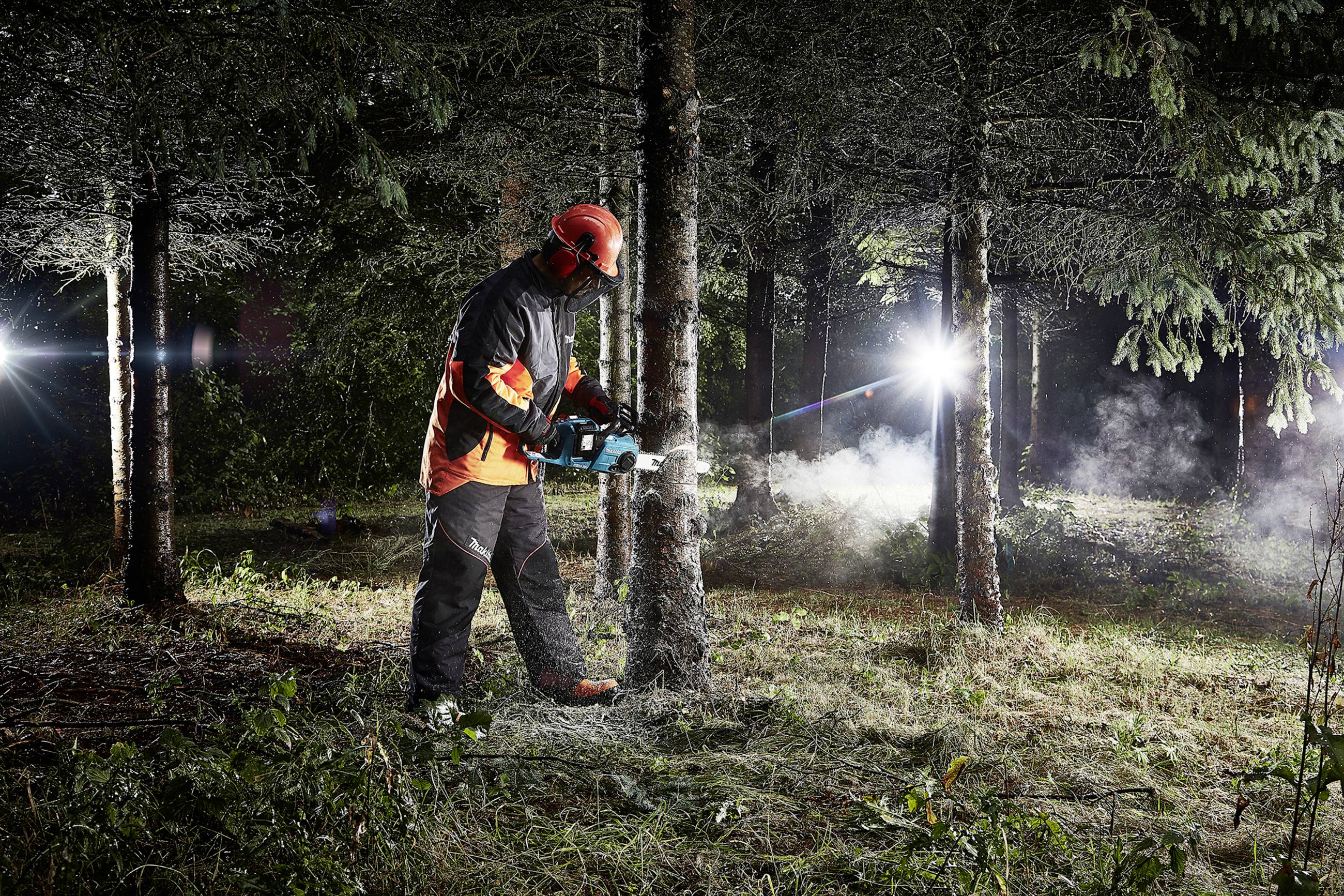 A forestry worker is sawing a tree with a chainsaw in a wooded area. He is wearing protective equipment and an orange safety helmet.
