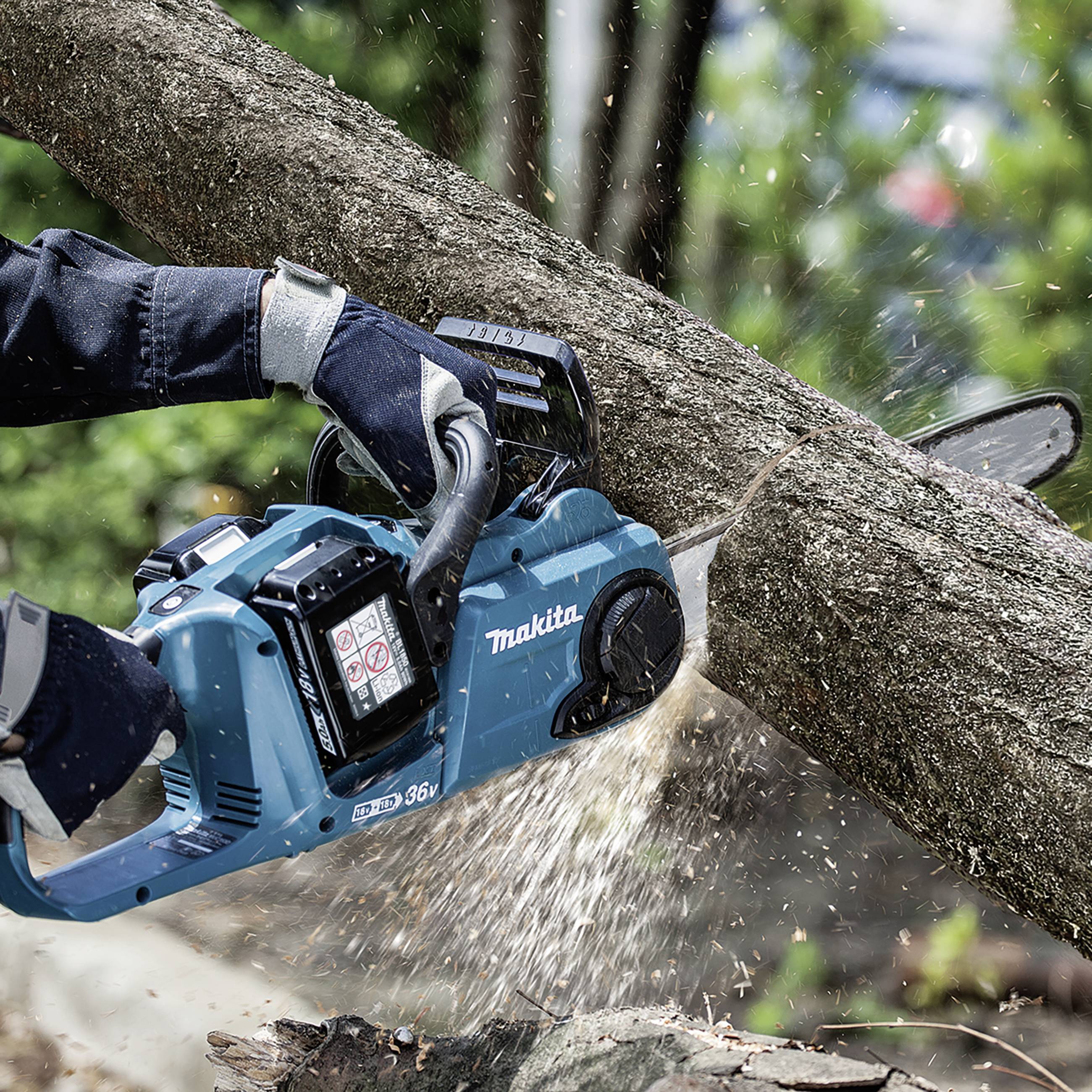 A worker is using a blue chainsaw to cut a thick, diagonally positioned branch in a wooded area.