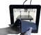 A 3D printer is printing an object made of plastic. Hands are holding a flexible printing plate with a safety notice.