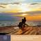 A person is fishing at sunset on a wooden pier by the sea, with an open fishing equipment box beside them.