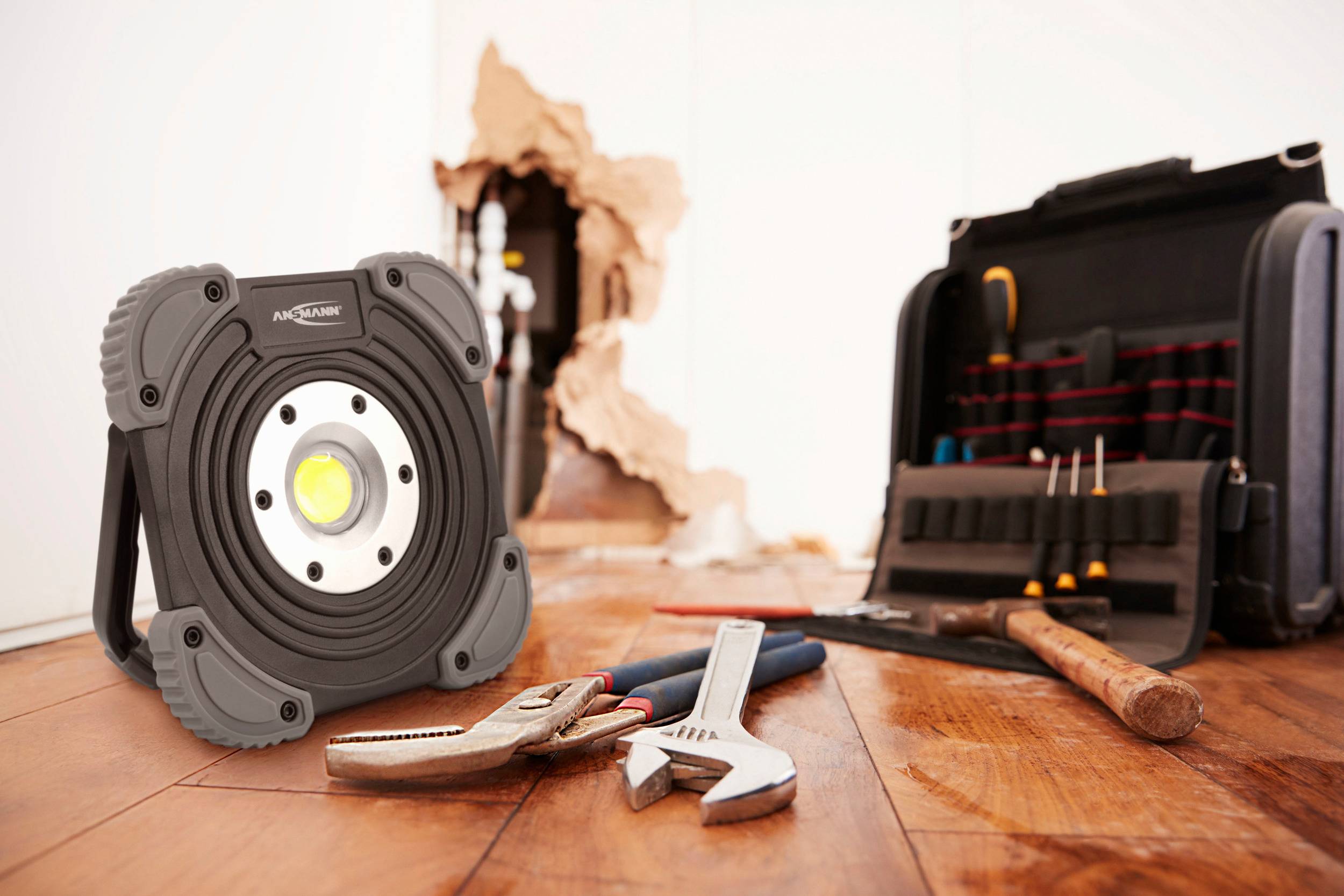 A construction site lamp stands on a wooden floor, surrounded by tools such as pliers and spanners. In the background, there is a hole in the wall.