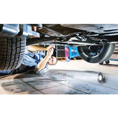 A person is repairing something underneath a car in a workshop. Tools and a work light are visible on the floor.