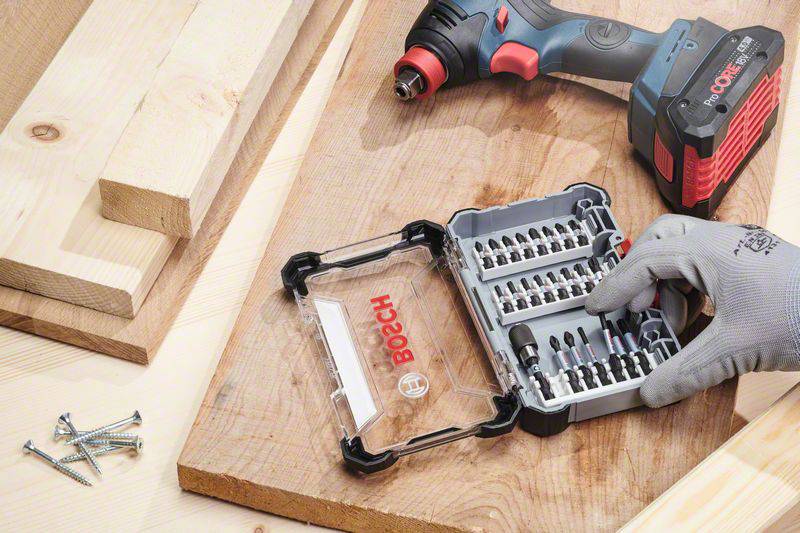 A cordless drill, Bosch screwdriver bit set and wooden planks on a workbench. A gloved hand is picking up a bit from the box.