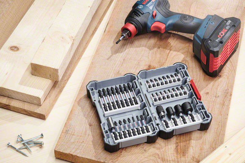 Cordless drill with an open case full of drill bits and driver bits on a wooden table, next to wooden planks and screws.