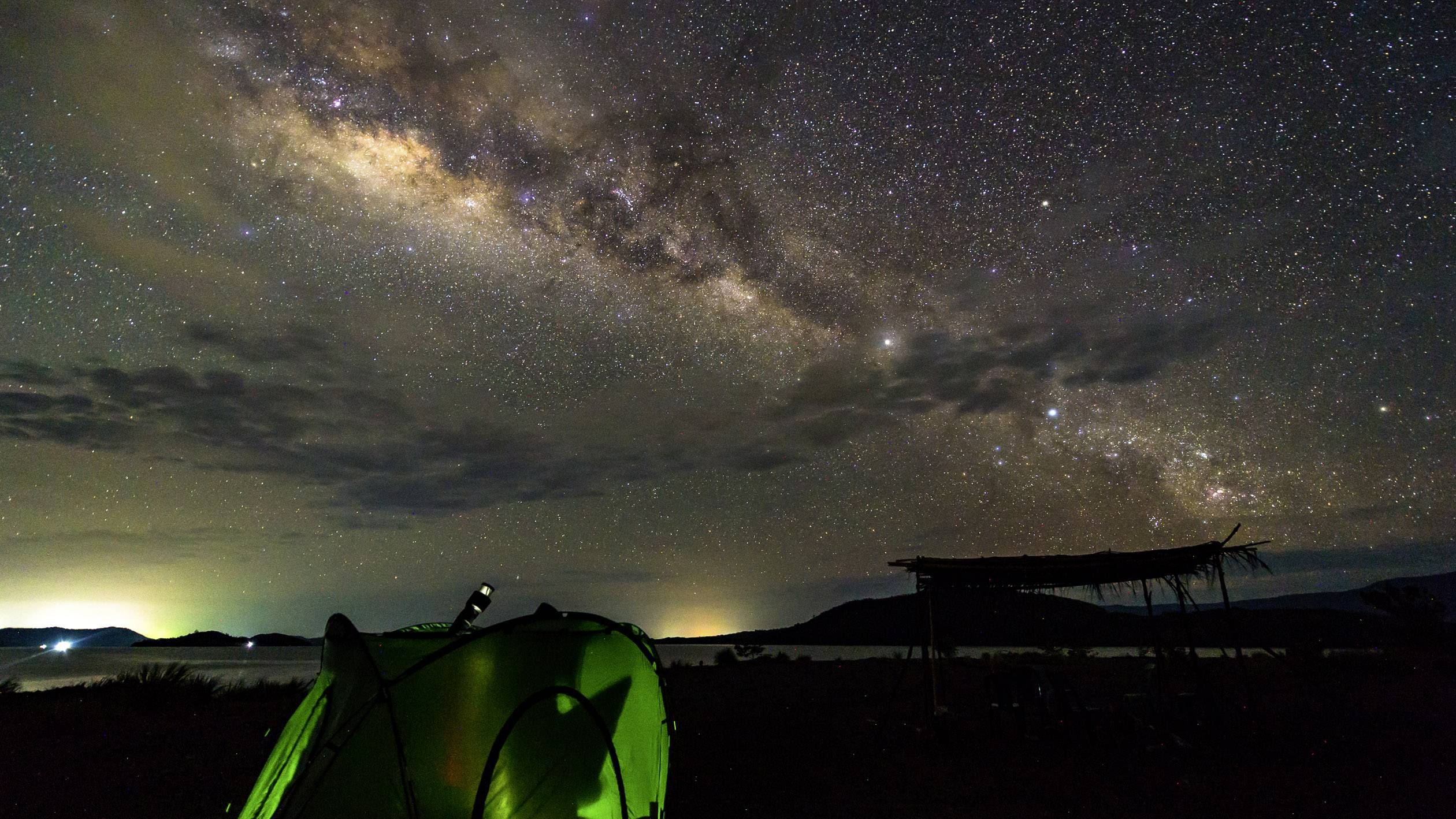 A green tent stands beneath a clear night sky, traversed by the Milky Way. Dark hills can be seen in the background.