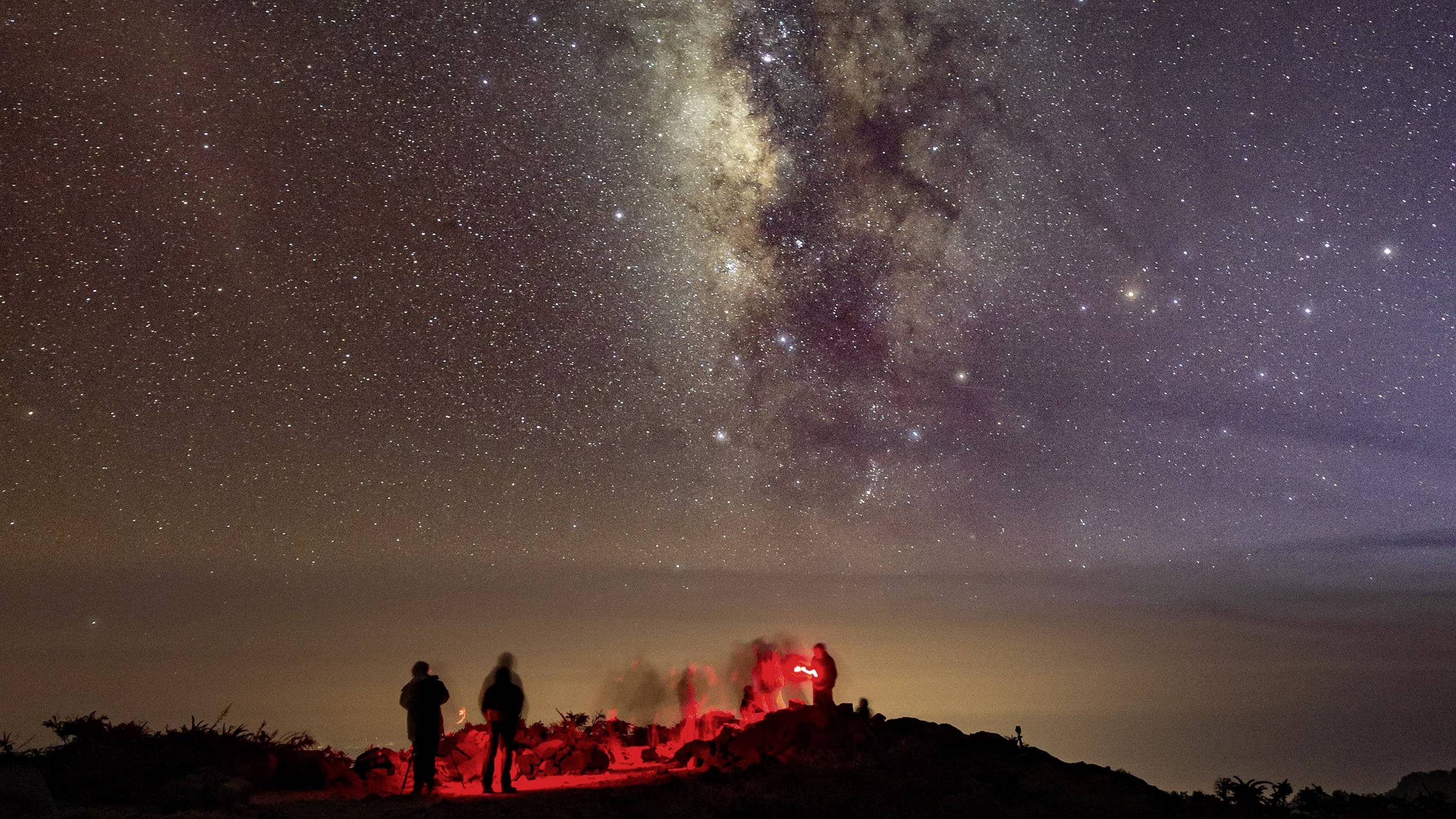 A group of people observes a star-filled night with the Milky Way in the background, red lights illuminating the ground.