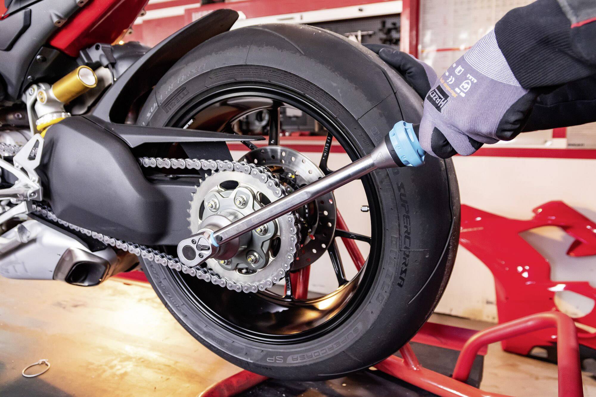 A tool is used to adjust the rear wheel of a motorbike. The technician is wearing gloves and working in a workshop environment.