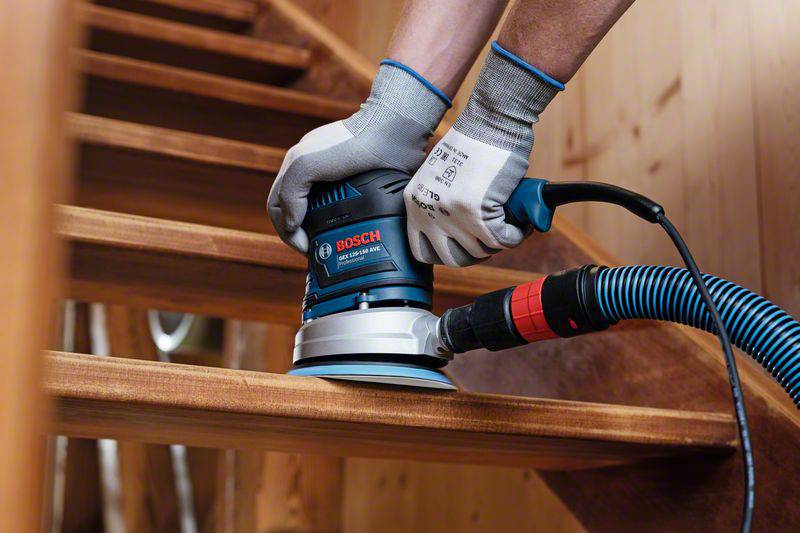 A person is sanding a wooden staircase with an electric sanding tool. They are wearing work gloves.