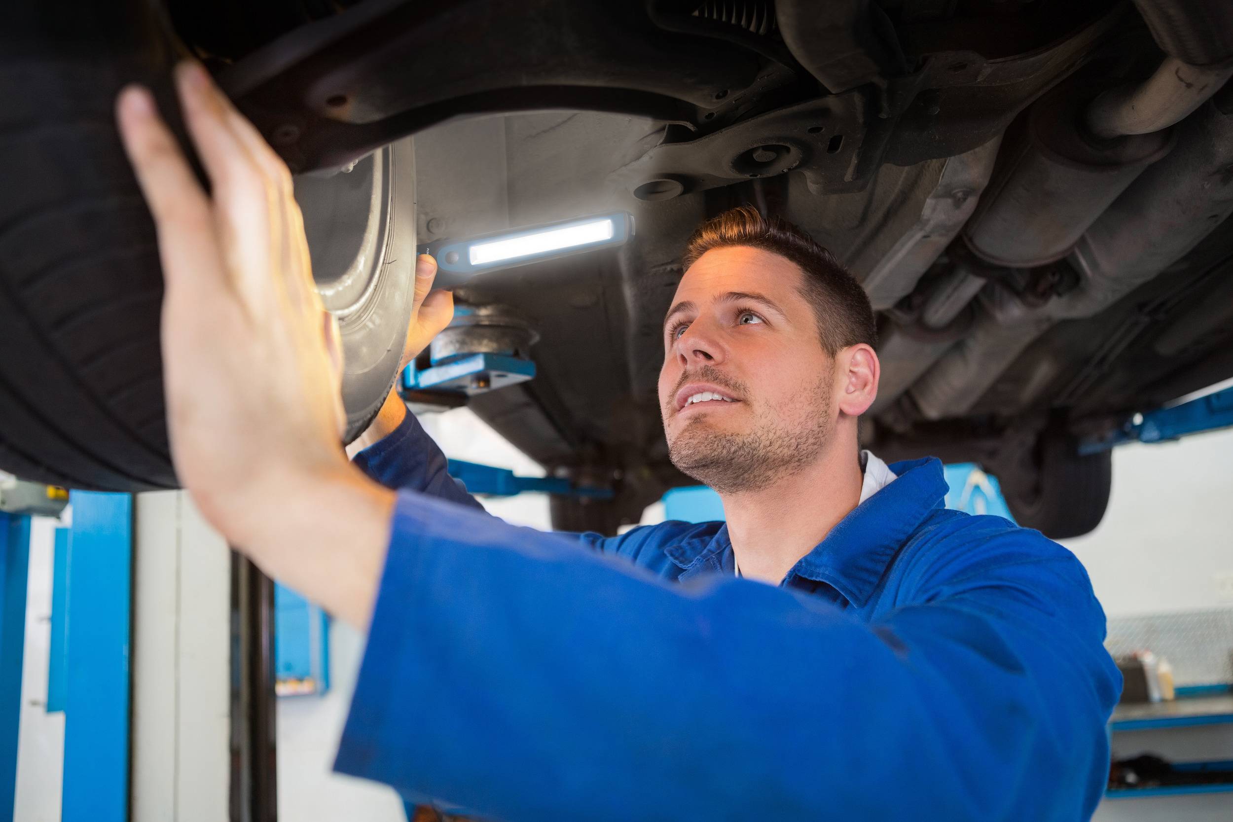 A mechanic in blue overalls is inspecting the underside of a car on a vehicle lift in a garage.