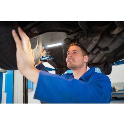A mechanic in blue overalls is inspecting the underside of a car on a vehicle lift in a garage.
