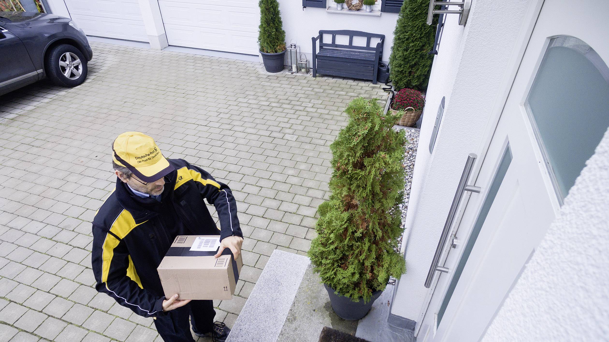 A delivery person in a yellow uniform is carrying a package to the front door of a house. In the background, a car and a closed garage can be seen.