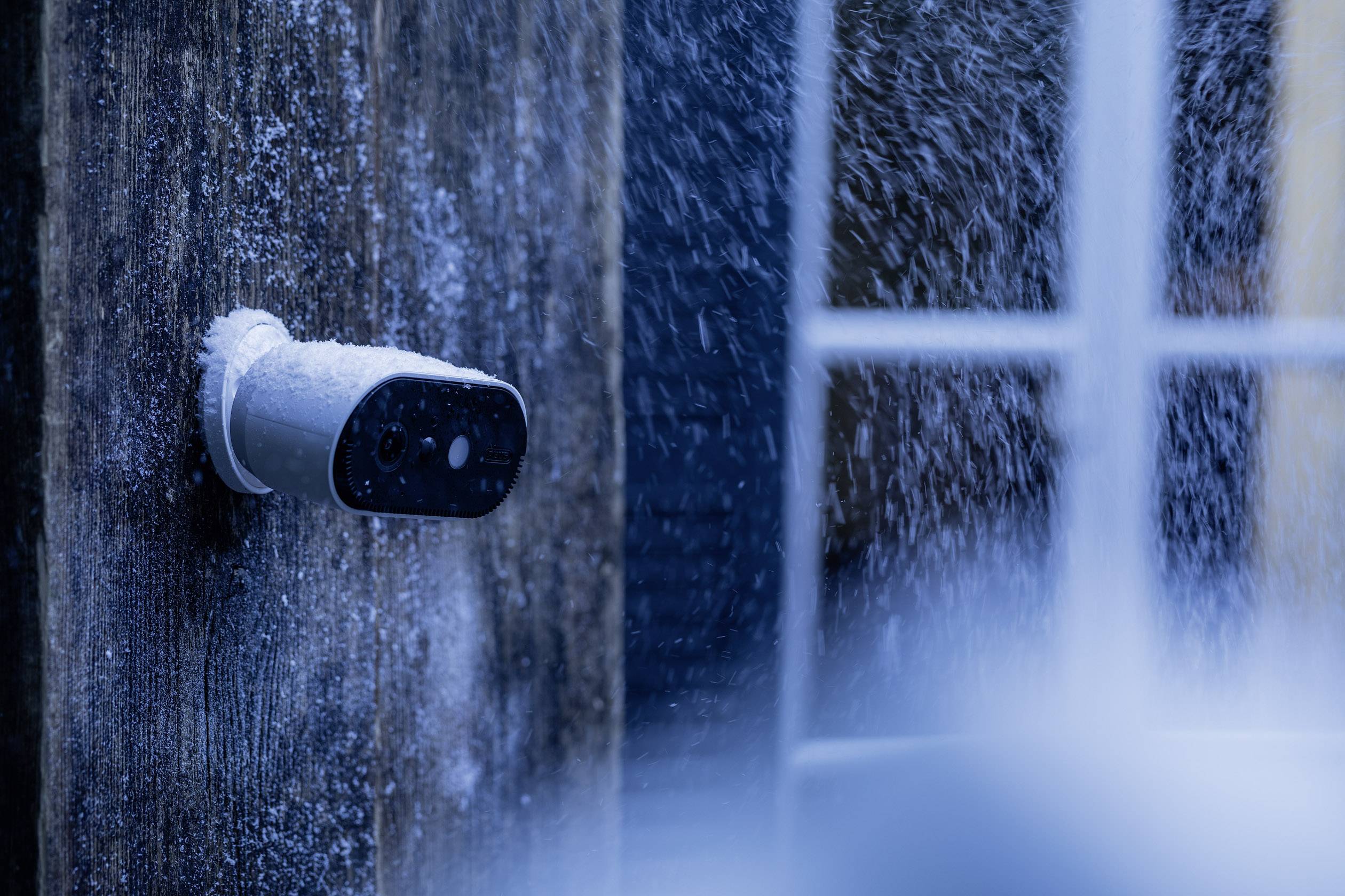 A snow-covered surveillance camera is mounted on a house wall, with snow falling in the background.
