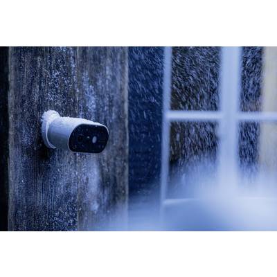 A snow-covered surveillance camera is mounted on a house wall, with snow falling in the background.