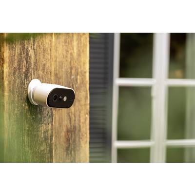 A security camera is mounted on the exterior wall of a house, with a window visible in the background.