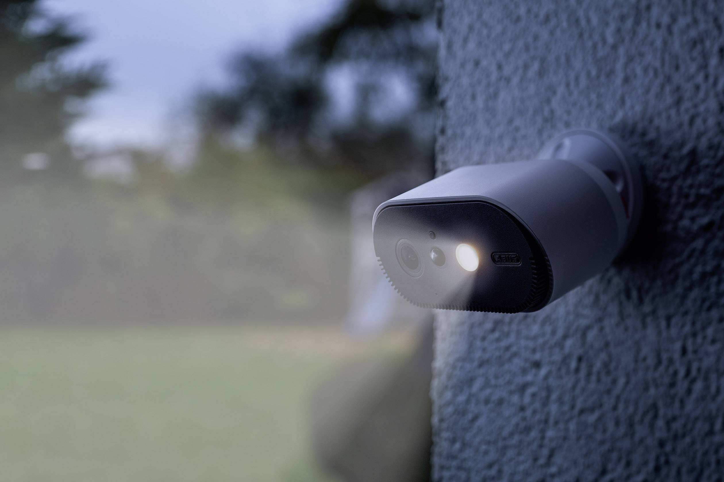 A fixed security camera mounted on a wall focused on a garden at dusk, with lights switched on.