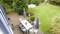 A terrace with garden furniture under parasols, surrounded by a green garden with a lawn and a wooden table, with a swing in the background.