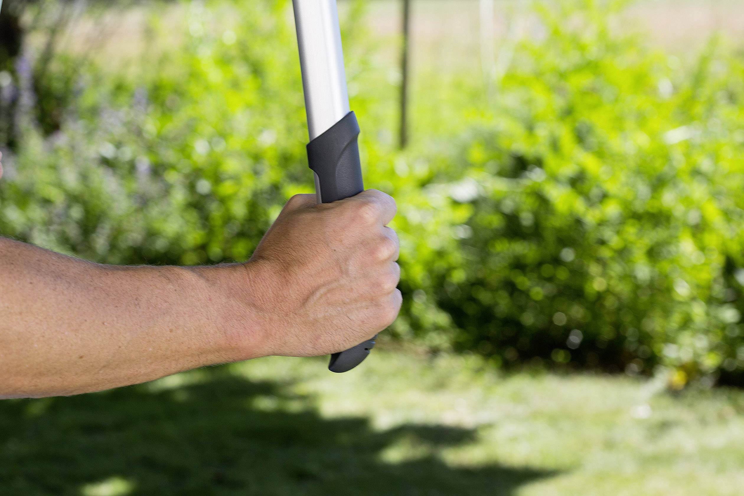 A hand grips a broom handle, with blurred green foliage visible in the background.