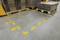 Yellow markings on the floor of a warehouse indicate a safe walking area. Pallets with cardboard boxes are positioned adjacent to the markings.