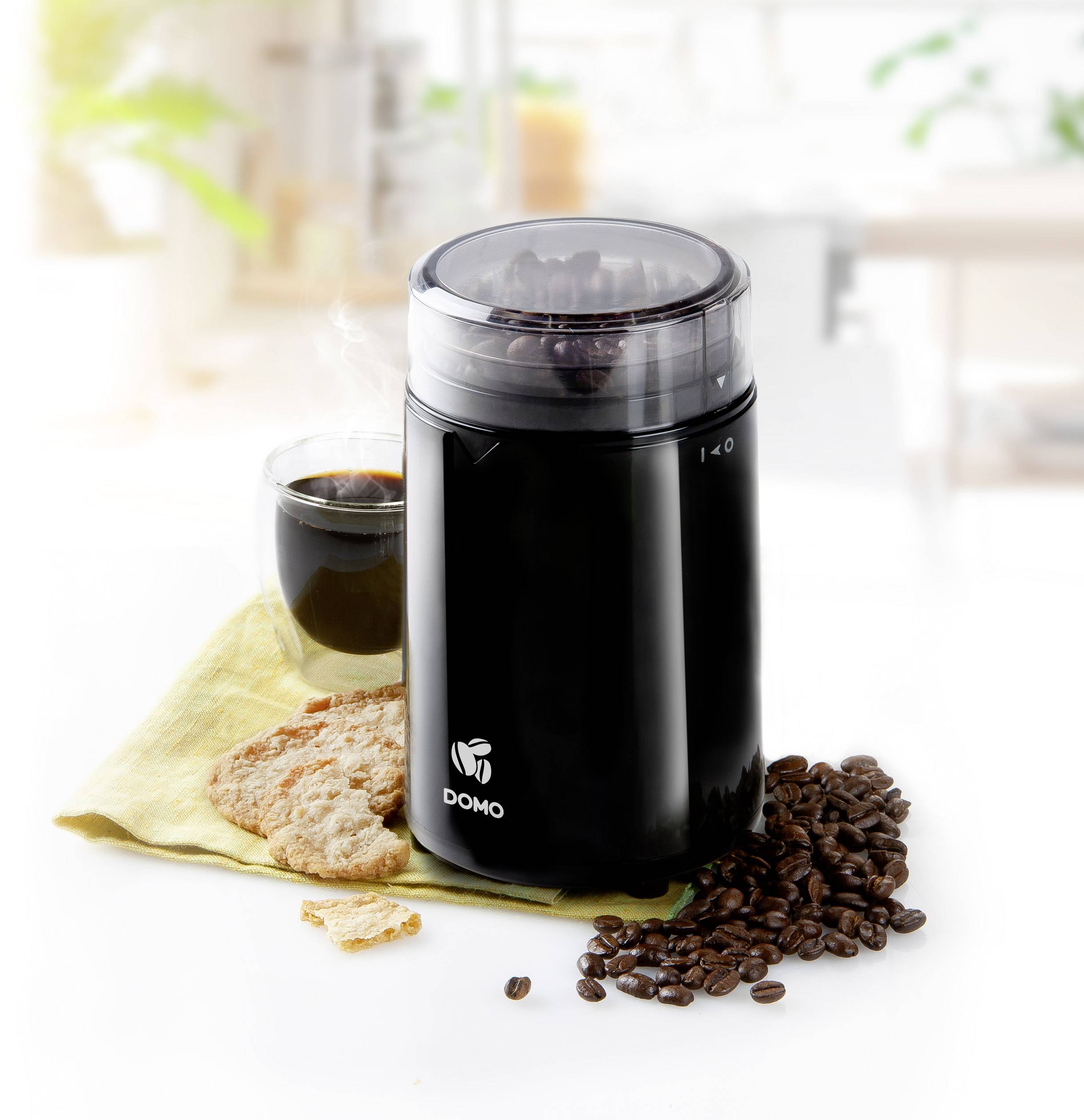 'Scene shows a black coffee grinder next to a coffee cup, biscuits and scattered coffee beans on a cloth in a bright kitchen.'