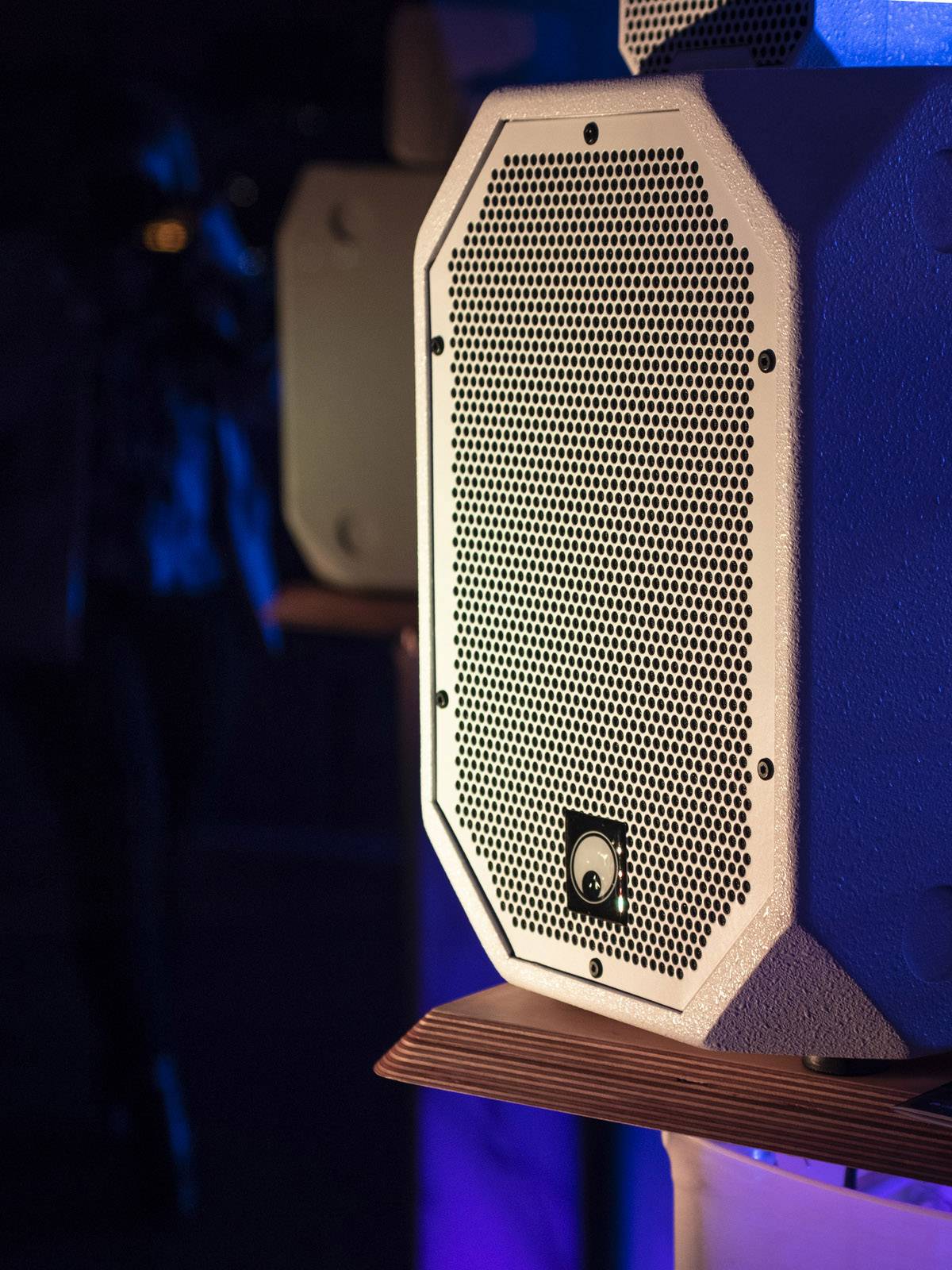 A large, white speaker with a grille at the front sits on a wooden table. The room is dimly lit.