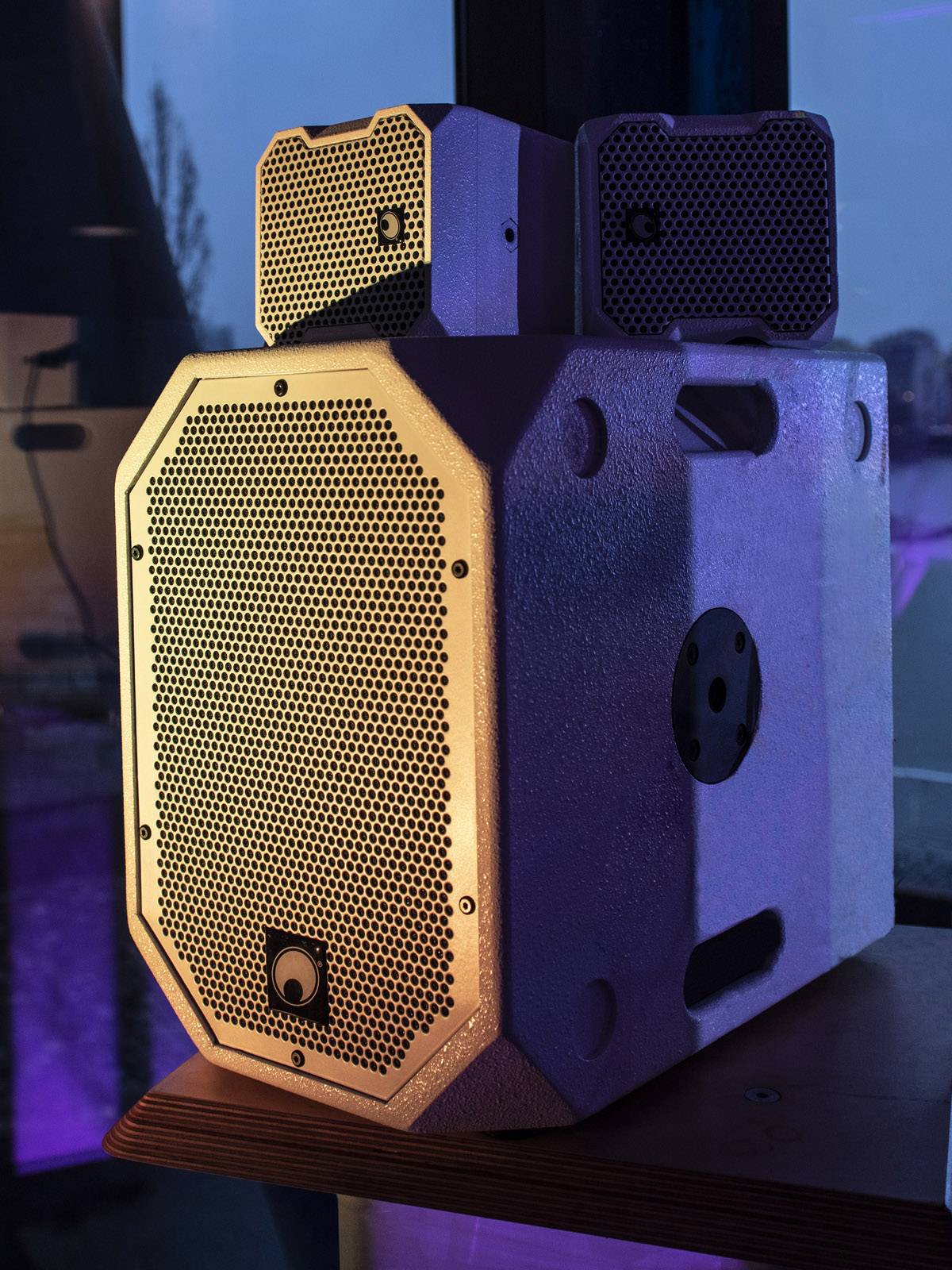 A grey speaker on a table, illuminated by blue and orange light, in a room with large windows in the background.