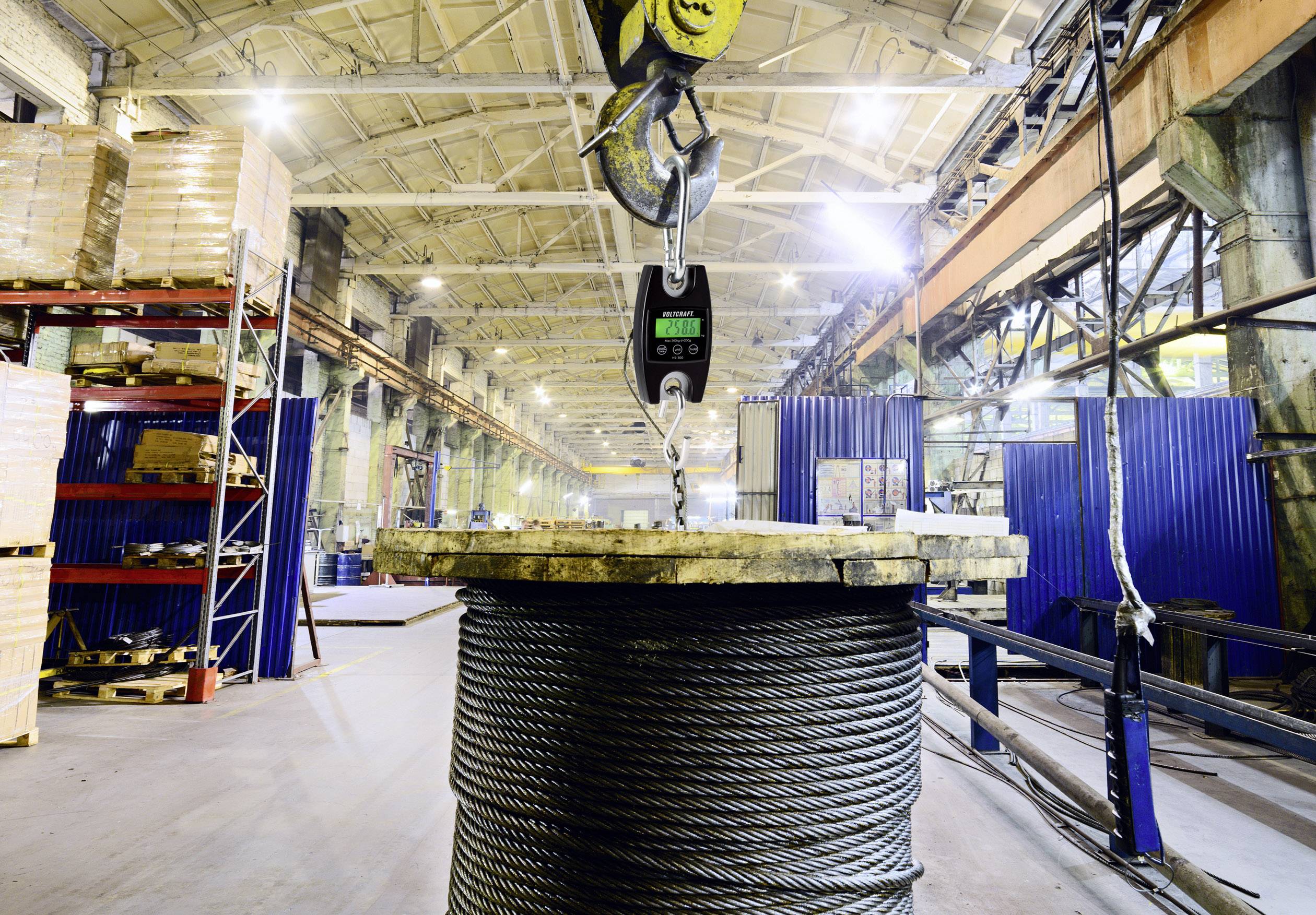 Large factory hall with a steel cable on a reel being lifted by a crane. Shelves and machinery are visible in the background.