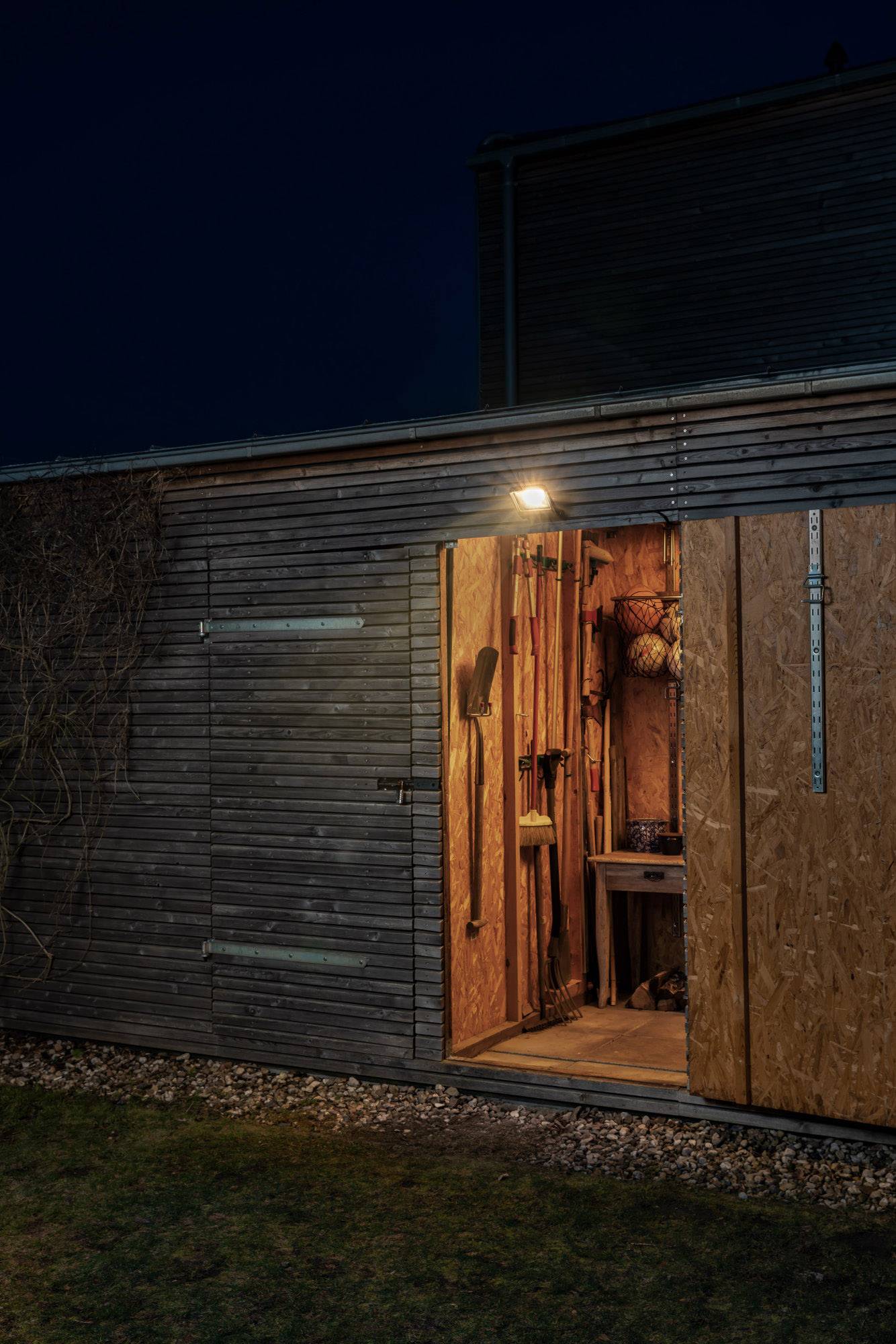 A shed at night with an open door, illuminated inside. Garden tools are visible.