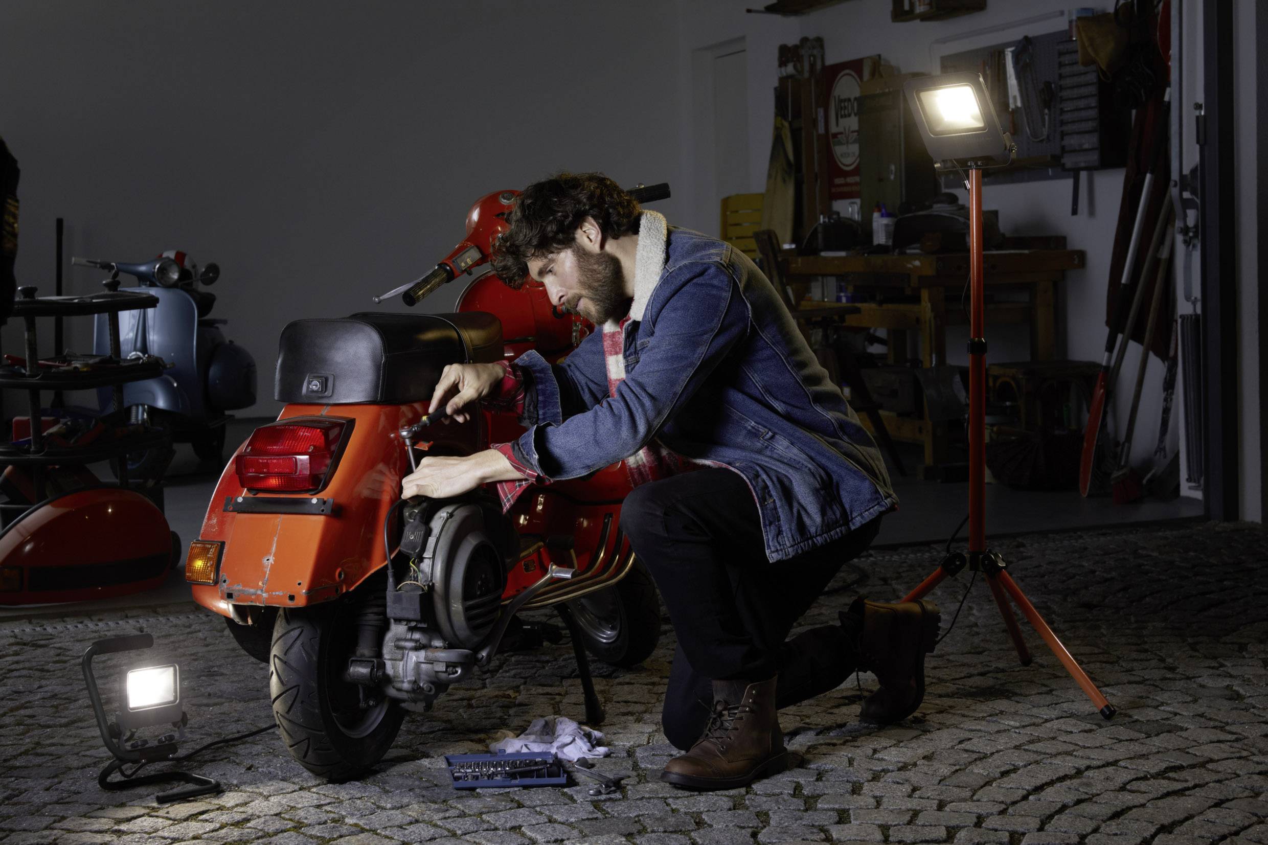 A man is repairing a red scooter in a garage. He is surrounded by work lights, wearing a denim jacket and focusing intently on the task.