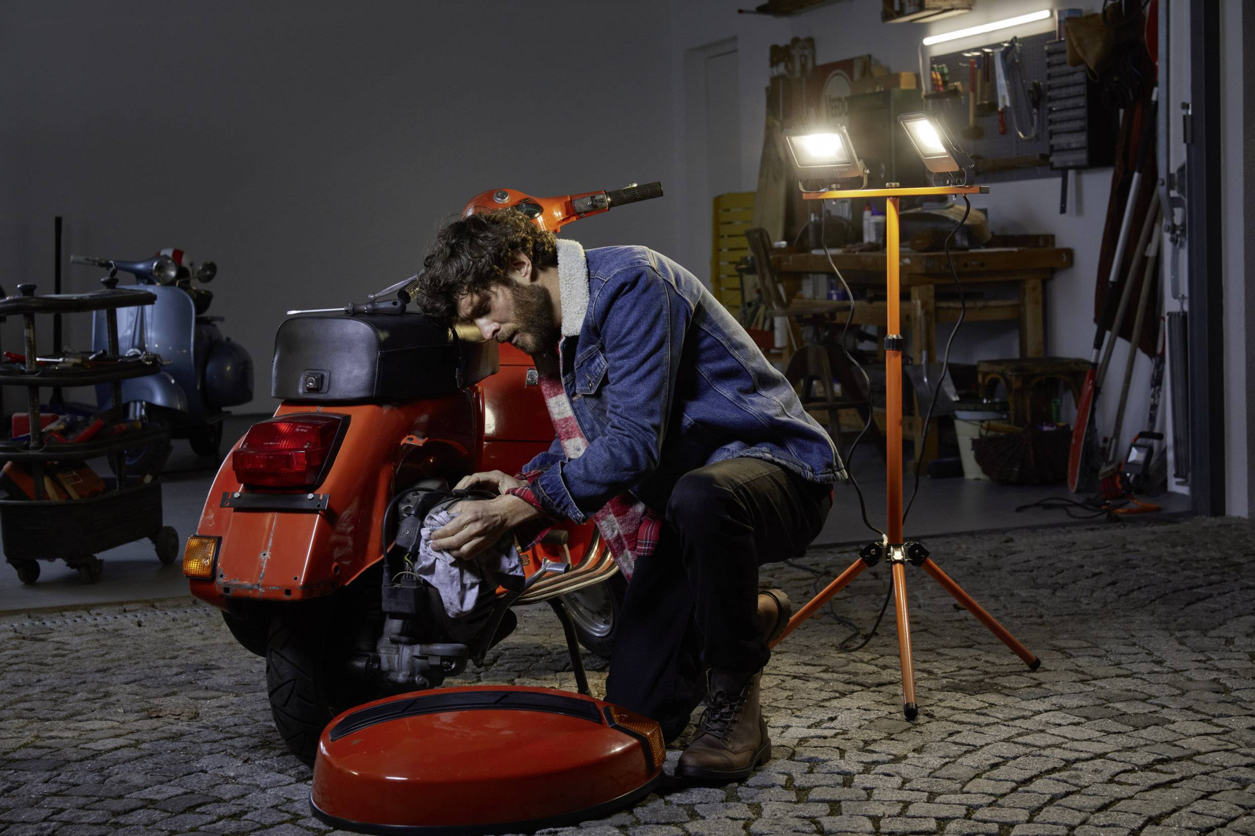 A man in a garage is repairing a red scooter. He is wearing a blue denim jacket and working intently under bright light.