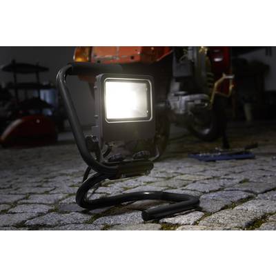 A construction floodlight illuminates a paved area in a workshop. A machine is blurrily visible in the background.