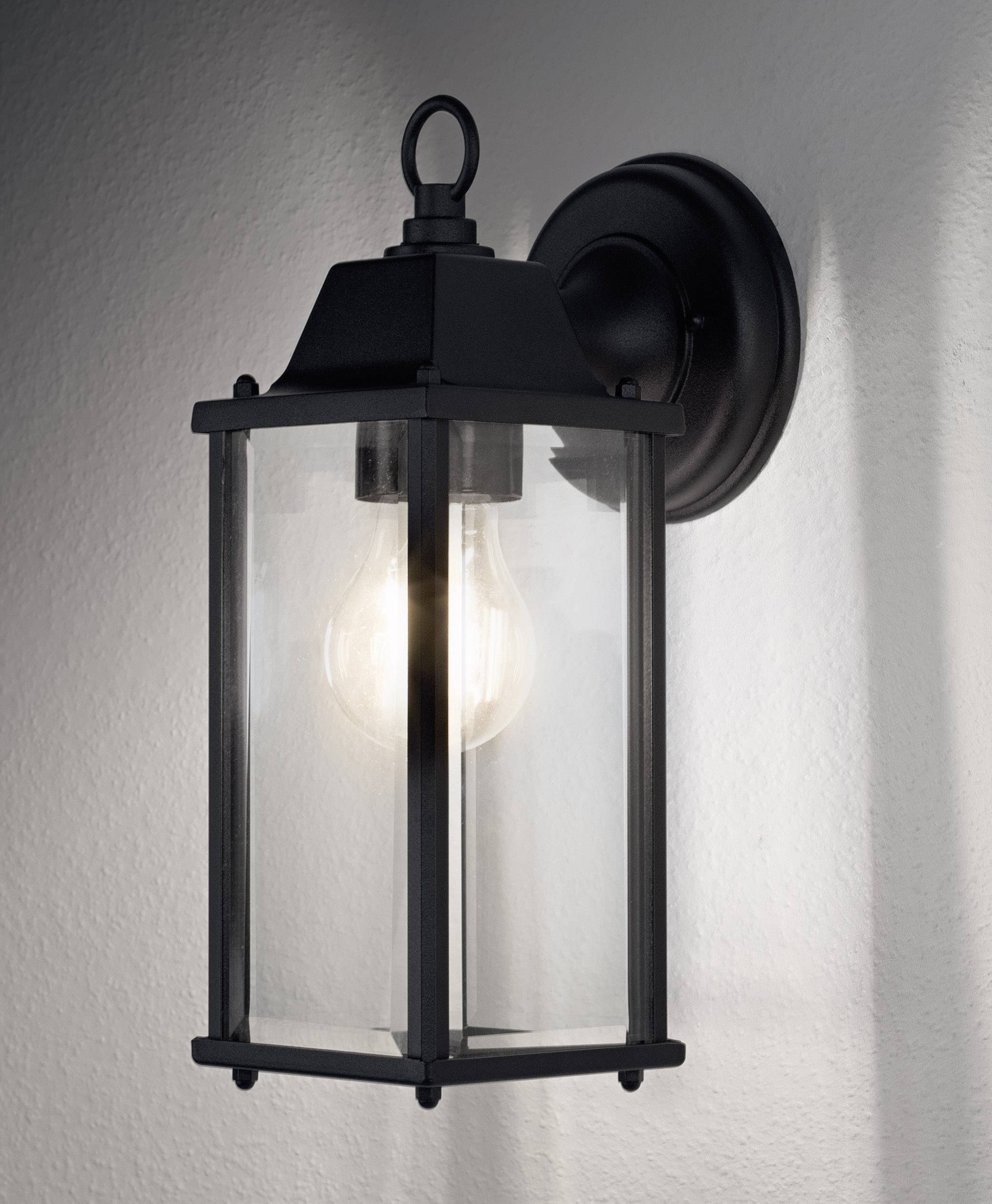 A black outdoor wall light with a glass body, radiating warm light. Mounted on a white wall, it provides illumination.