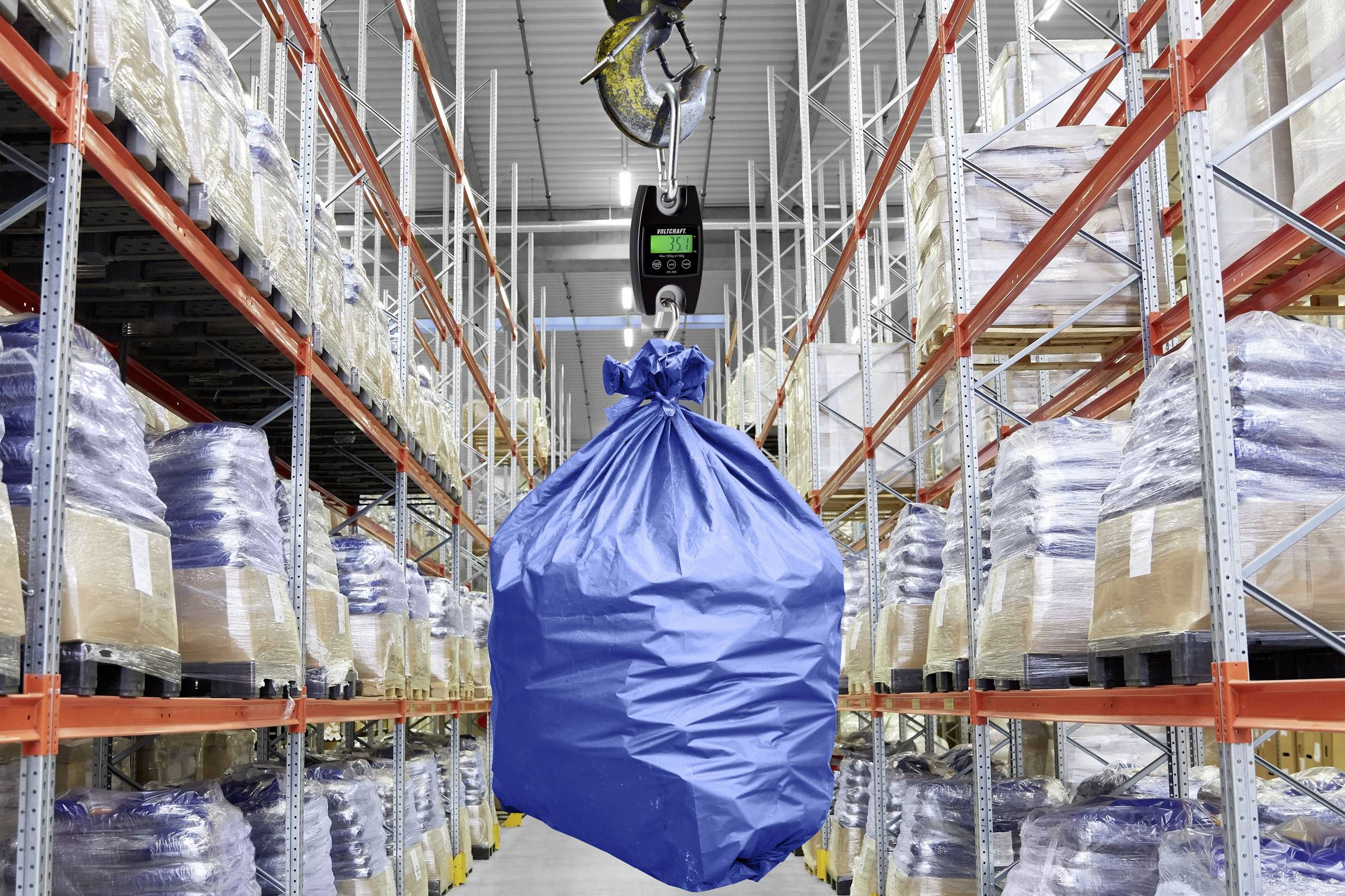A blue bag is being lifted by a crane with a digital scale in a warehouse. Shelves with cardboard boxes and sacks are visible in the background.