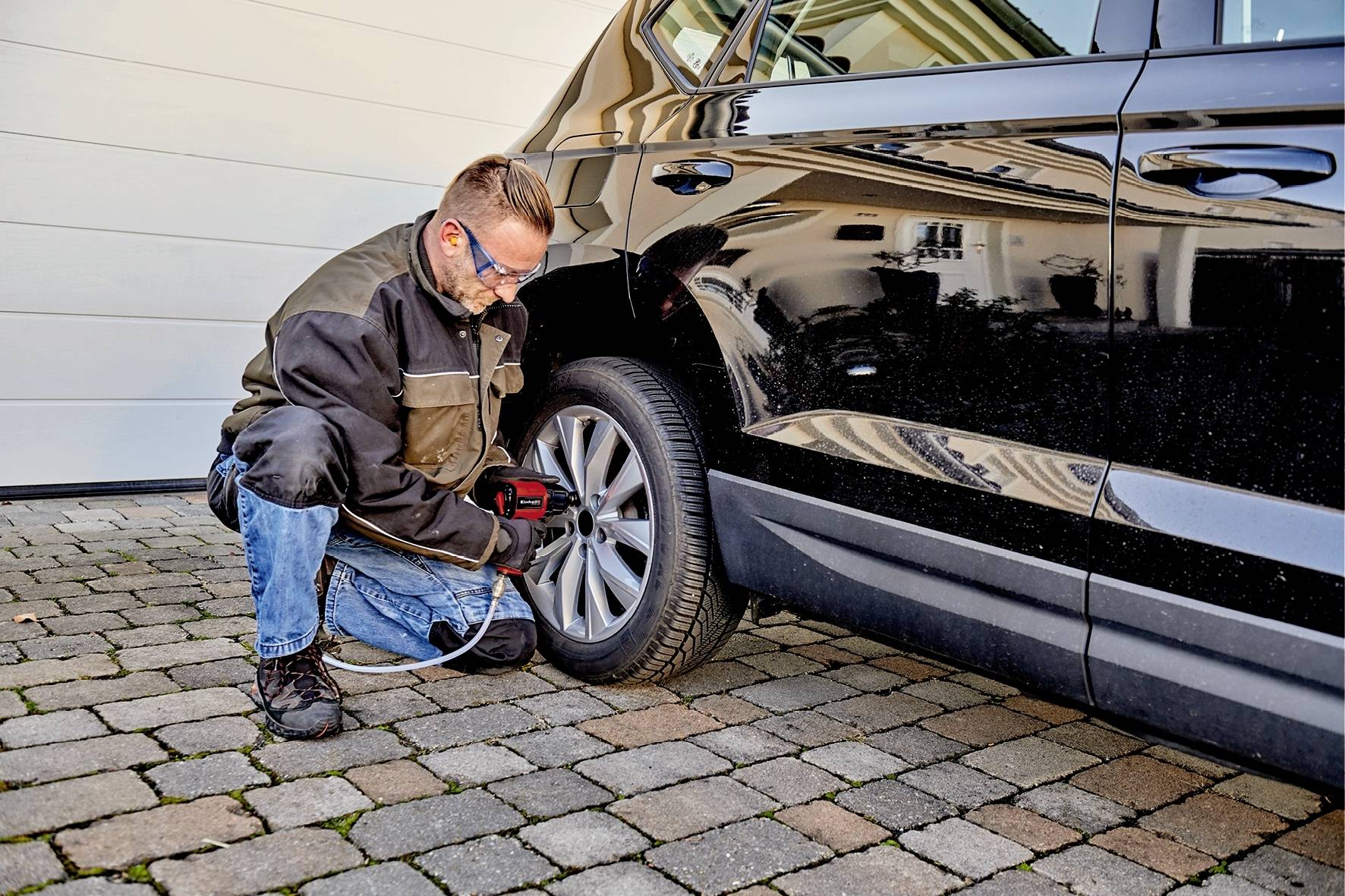 A man is changing the tyre of a black car in a driveway. He is wearing work clothes and using a tool to loosen the wheel nuts.