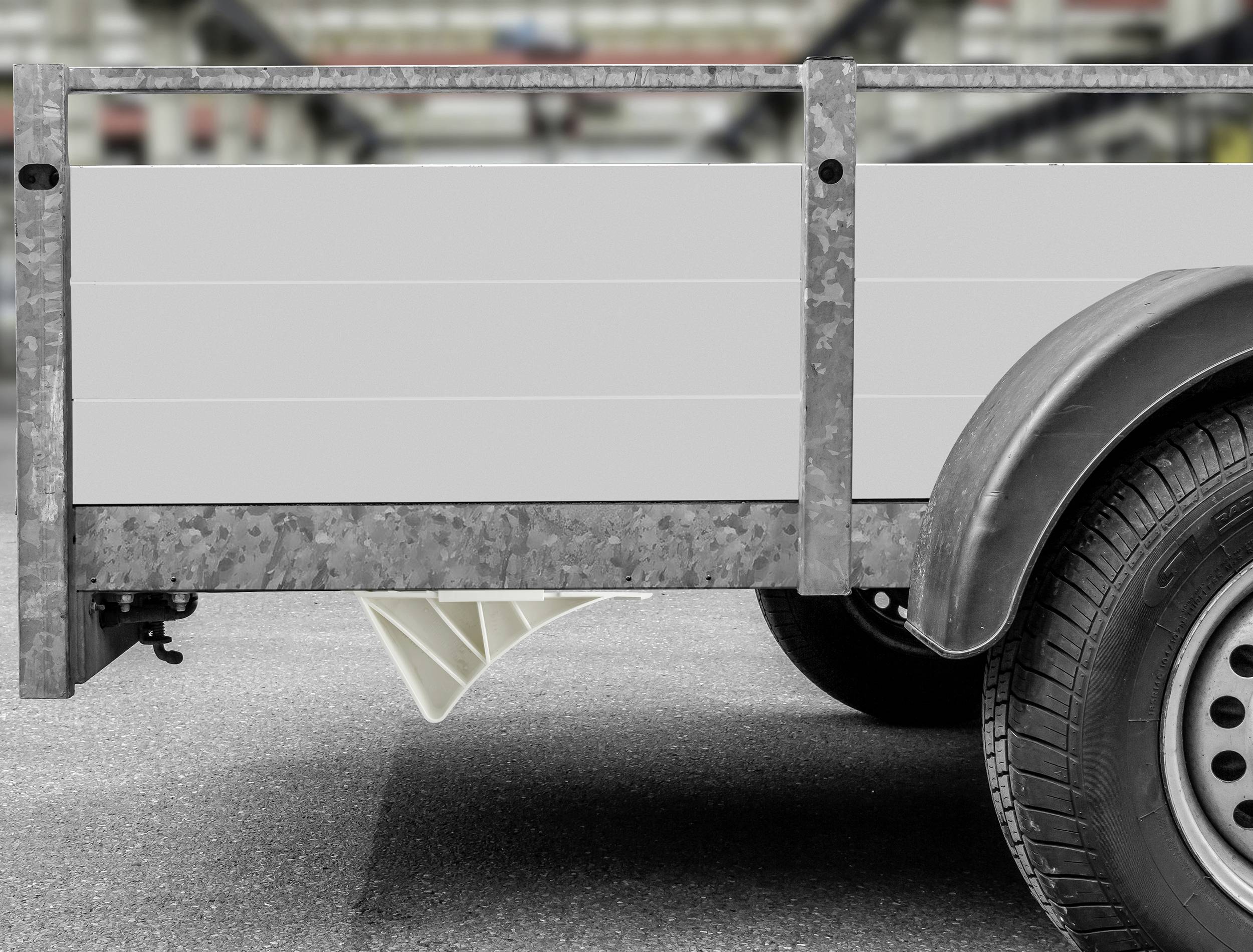 A grey trailer on a road, with a focus on the wheel and side walls of the trailer. A blurry urban environment is visible in the background.