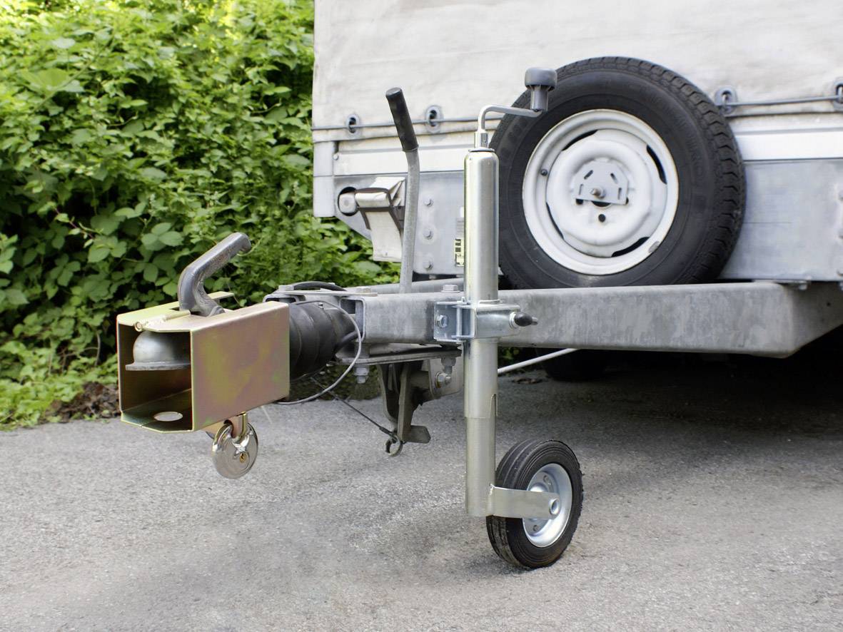 Trailer drawbar with jockey wheel and spare wheel fitted, supported on an asphalt surface with a green hedge in the background.