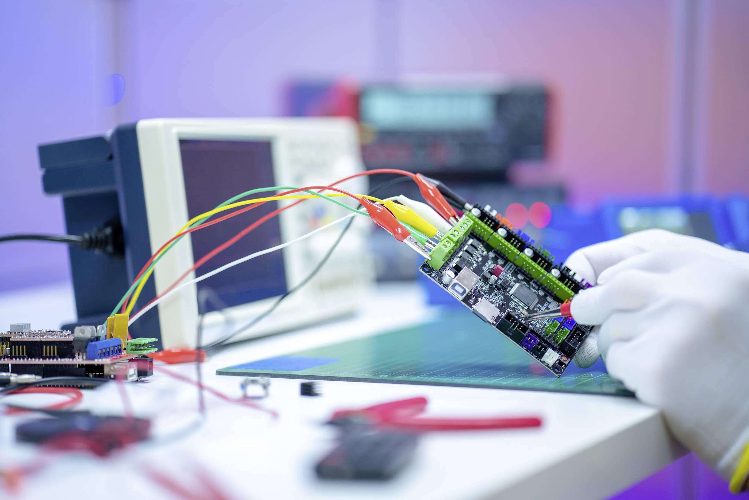 A close-up shows a person wearing gloves holding an electronic circuit board with several connected cables. Measuring instruments are visible in the background.